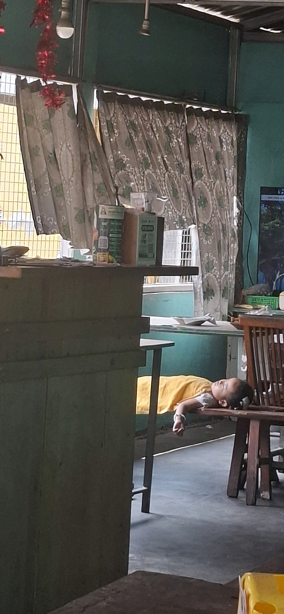 A child is seen sleeping on a wooden chair in an indoor setting. The child is wearing a yellow shirt and appears to be in a relaxed sleeping posture, with their head resting on the armrest or seat of the chair. The background shows a room with green walls, curtains with a patterned design, and a shelf with some boxes. A television screen is visible on the right side of the image. The floor is a light grey color. The lighting suggests it is daytime. The overall scene appears to be a domestic or casual environment, possibly a home or a shop.