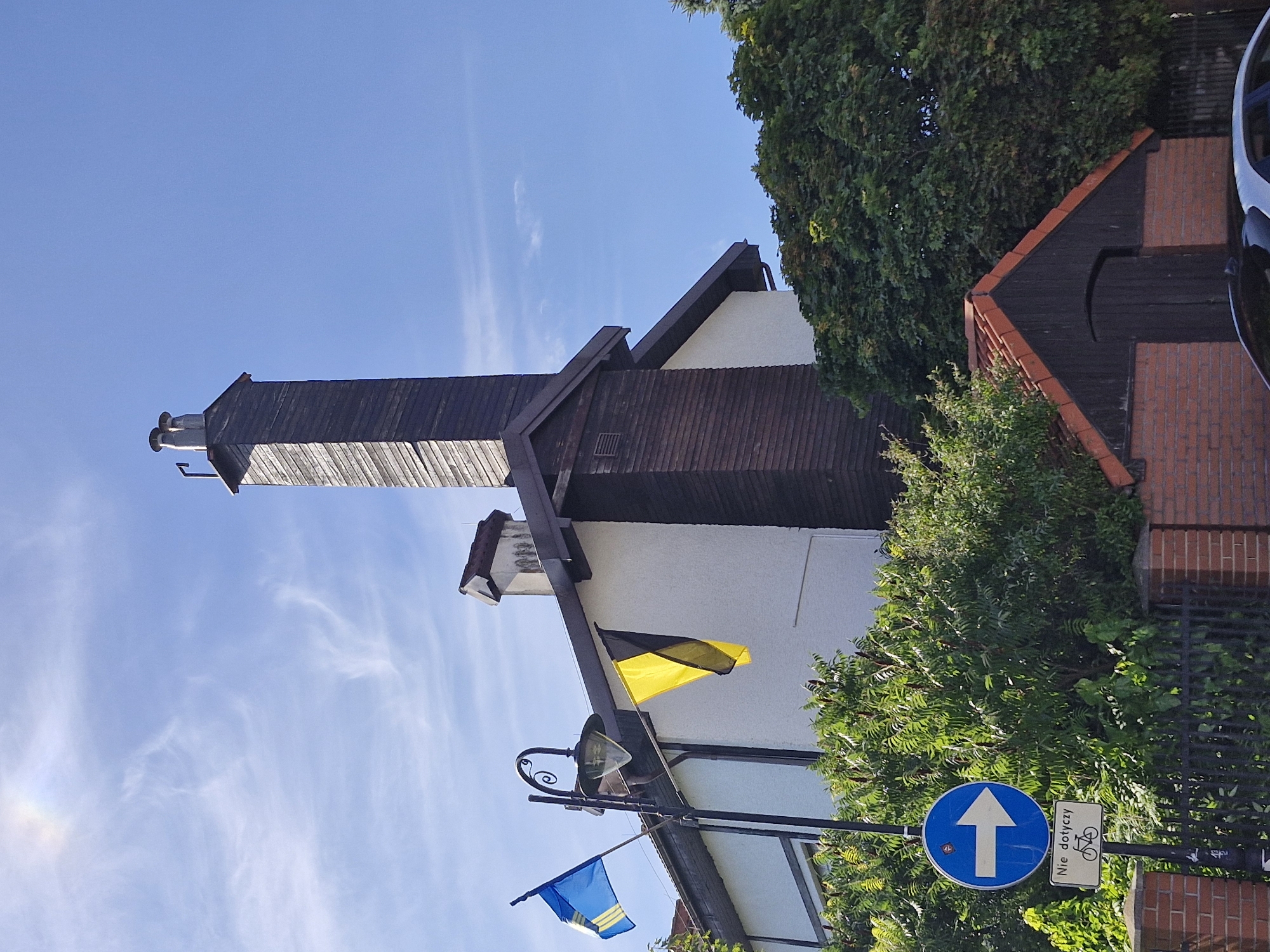 The image captures a building in Hel, Poland, with a prominent wooden chimney and two flags displayed on poles outside. One flag is blue and yellow, resembling the Ukrainian flag. The other flag is black and yellow, possibly a regional or historical flag. The building appears to be a house with a white facade and dark brown accents. In front of the building, dense green foliage obscures a brick wall and fence. A street sign with a white arrow pointing to the right is visible, along with a smaller sign below it depicting a bicycle and text that reads "Nie dotyczy," meaning "Does not apply" in Polish, indicating that the preceding sign's instruction does not apply to cyclists. The sky is blue with some wispy clouds, and a faint rainbow is visible in the upper left corner. A small, red-tiled shed with a dark wooden door is also present in the foreground. The scene suggests an outdoor setting in a residential or commercial area.