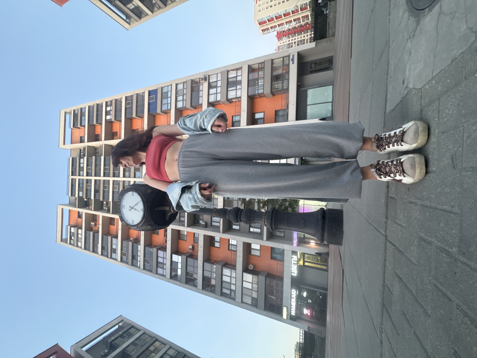The image depicts a woman standing next to a black street clock in an urban environment. The woman is wearing a red top, gray pants, a light blue denim jacket, and white sneakers with leopard print laces. She is looking down at the clock. The clock face is white with black numerals and hands. Behind the woman and the clock are modern, high-rise apartment buildings, featuring a combination of gray, orange, and white facades. The lower levels of the buildings appear to house commercial spaces, with visible storefronts displaying signs with Russian text. The sky is a pale blue. The location appears to be in Tyumen, Russia, suggested by the signs visible on the buildings. The ground is paved with gray concrete blocks.