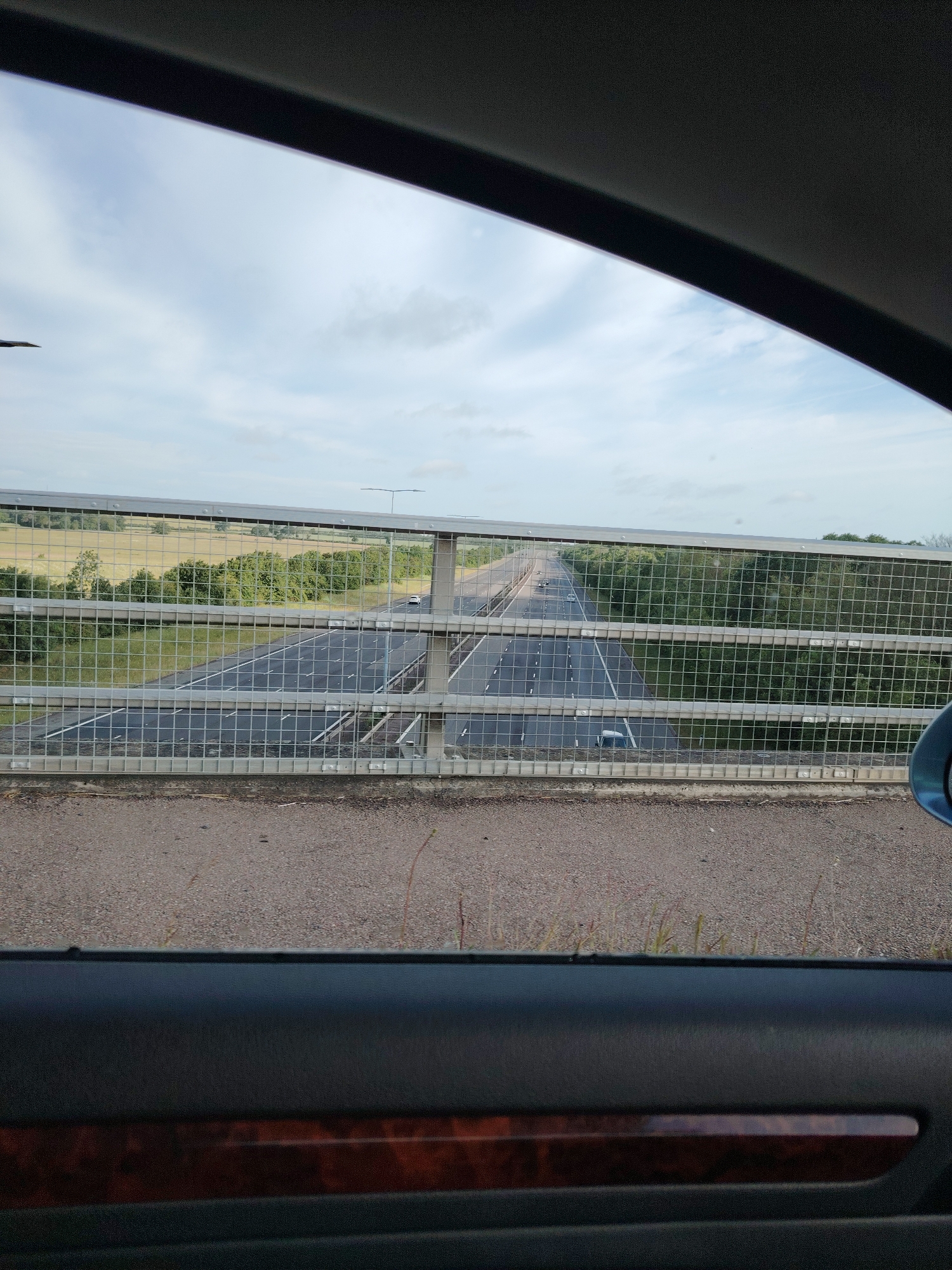The image captures a view from inside a vehicle, looking through a side window over a multi-lane highway from an elevated position, consistent with a bridge overpass in the Huntingdonshire region of the United Kingdom. The top section of the image shows a pale blue sky with scattered white and light grey clouds, framed by the dark interior trim of the vehicle's roofline.
The middle section of the image is dominated by a grey metal mesh barrier, typical of bridge safety fencing, positioned directly in front of the viewer. Through and beyond this barrier, a dual carriageway highway is visible, featuring at least two lanes of asphalt roadway in each direction, marked with white dashed and solid lines. Several vehicles are present on the highway; a white car and a darker vehicle are discernible in the lanes furthest from the viewer, while other vehicles appear further down the road. A central reservation, densely covered with green trees and bushes, separates the opposing directions of traffic. A single lamppost with a dark pole extends upwards from the right side of the central reservation.
The lower section of the image reveals the interior of the vehicle, specifically a dark grey car door panel. A polished wooden trim piece, rectangular in shape, is embedded horizontally within the door panel. Above the car door, a strip of grey, textured asphalt or concrete forms the surface of the bridge, with sparse blades of grass or weeds emerging from cracks. A small portion of a blue object, likely a side mirror, is visible on the far right edge of the window view.