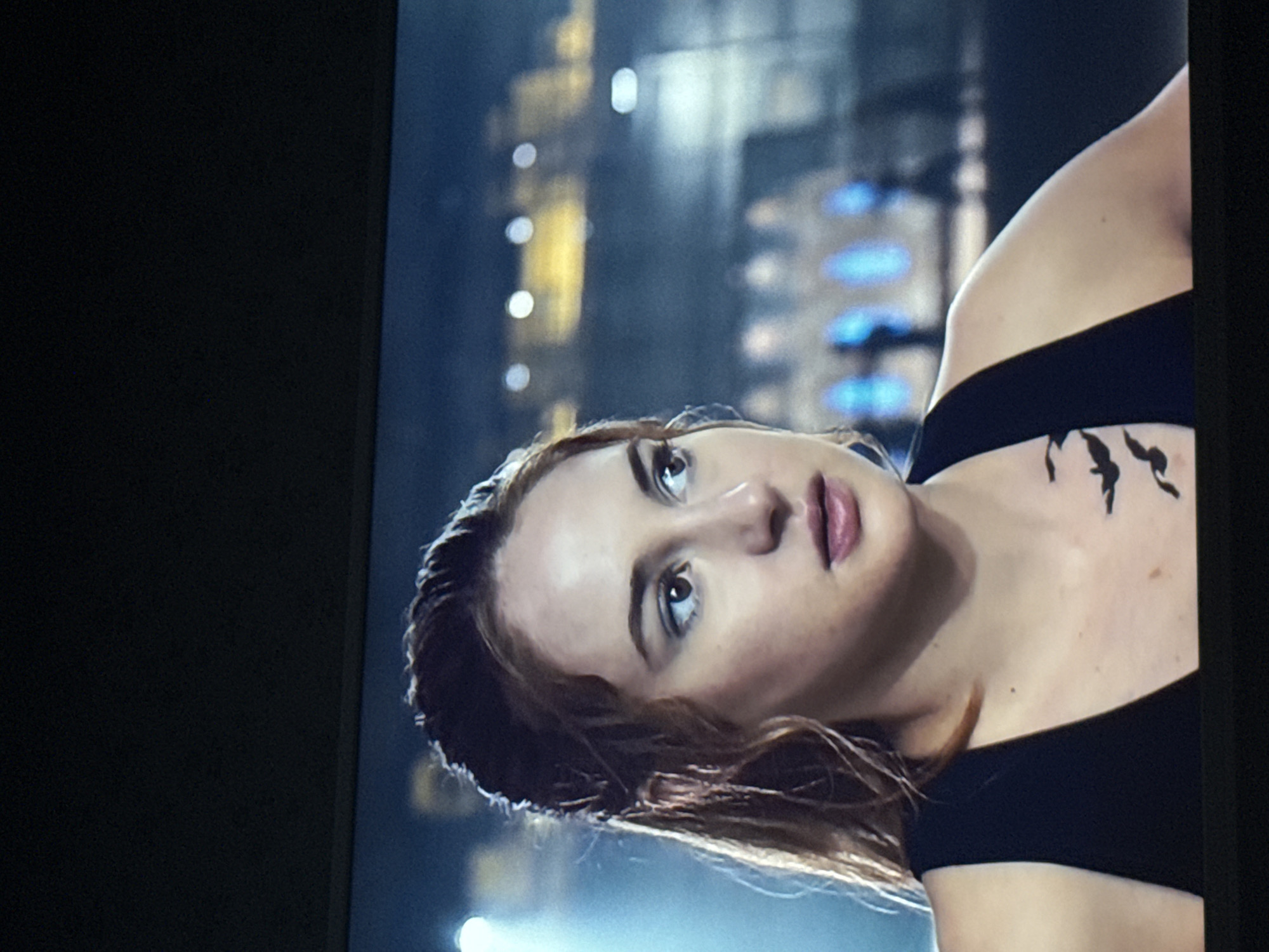 A close-up shot depicts a light-skinned woman with brown hair, possibly in her late teens or early twenties. Her gaze is directed upward and slightly to the left. She has a tattoo of three birds on her upper chest, near her shoulder. She is wearing a black top. The background is blurry, suggestive of a city at night, with out-of-focus lights and structures. There is no definitive evidence to identify the exact location as Mytishchi, Russia.