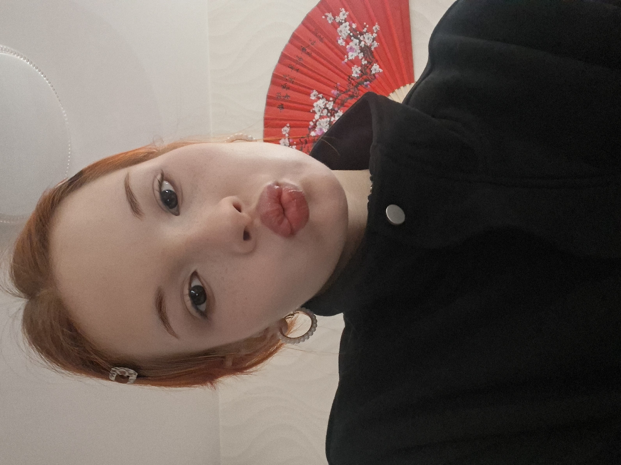 A close-up, tilted selfie features a young person with light red or auburn hair, fair skin, and scattered freckles on their nose and cheeks. The individual is making a playful "kissing face" with puckered lips and looking directly at the viewer with dark, expressive eyes. They have medium-thickness, well-groomed eyebrows.
The person is wearing a black jacket or top, evidenced by the visible fabric and a silver button on the right side of the frame. Decorative earrings are present: a small, clear or silver-toned clip-on style is visible in their hair above the left ear, and a larger, circular hoop earring with a textured or beaded edge adorns the right earlobe.
Behind the person's head, a vibrant red hand fan, partially fanned open, is prominently displayed. It features a decorative white cherry blossom (sakura) design. The background is a light-colored, plain wall, with a faint circular pattern or object, possibly a decorative panel or headboard, visible in the upper left.
The lighting is bright and even, indicating an indoor setting. The overall impression is casual and cheerful. No visible text is present in the image. The scene is set in Chaikovsky, Russia.