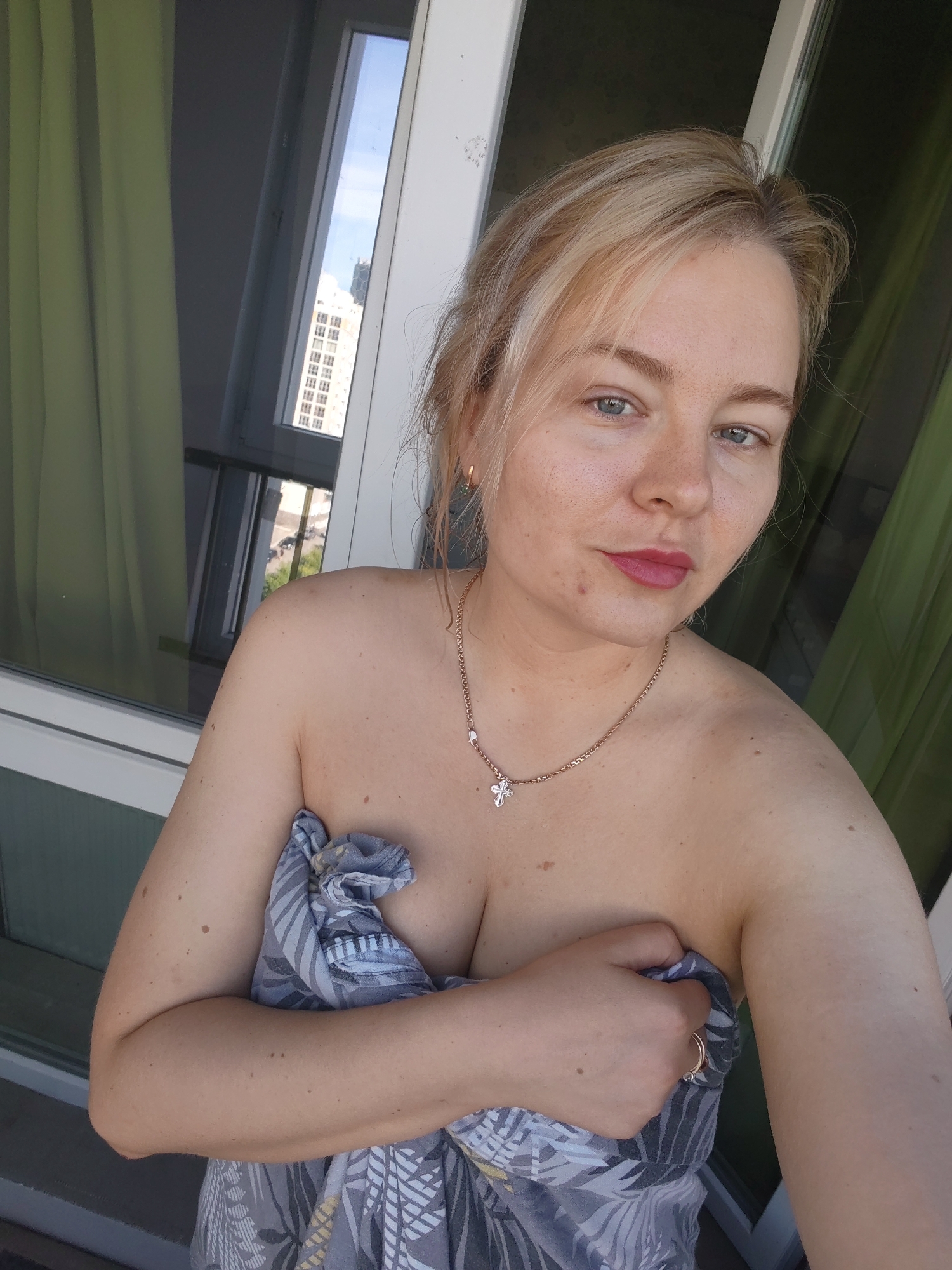 A blonde woman stands on a balcony in Ufa, Russia, holding a patterned towel to her chest. She wears a gold chain with a cross pendant and a gold ring on her right hand. The background shows a window with green curtains and a distant building.