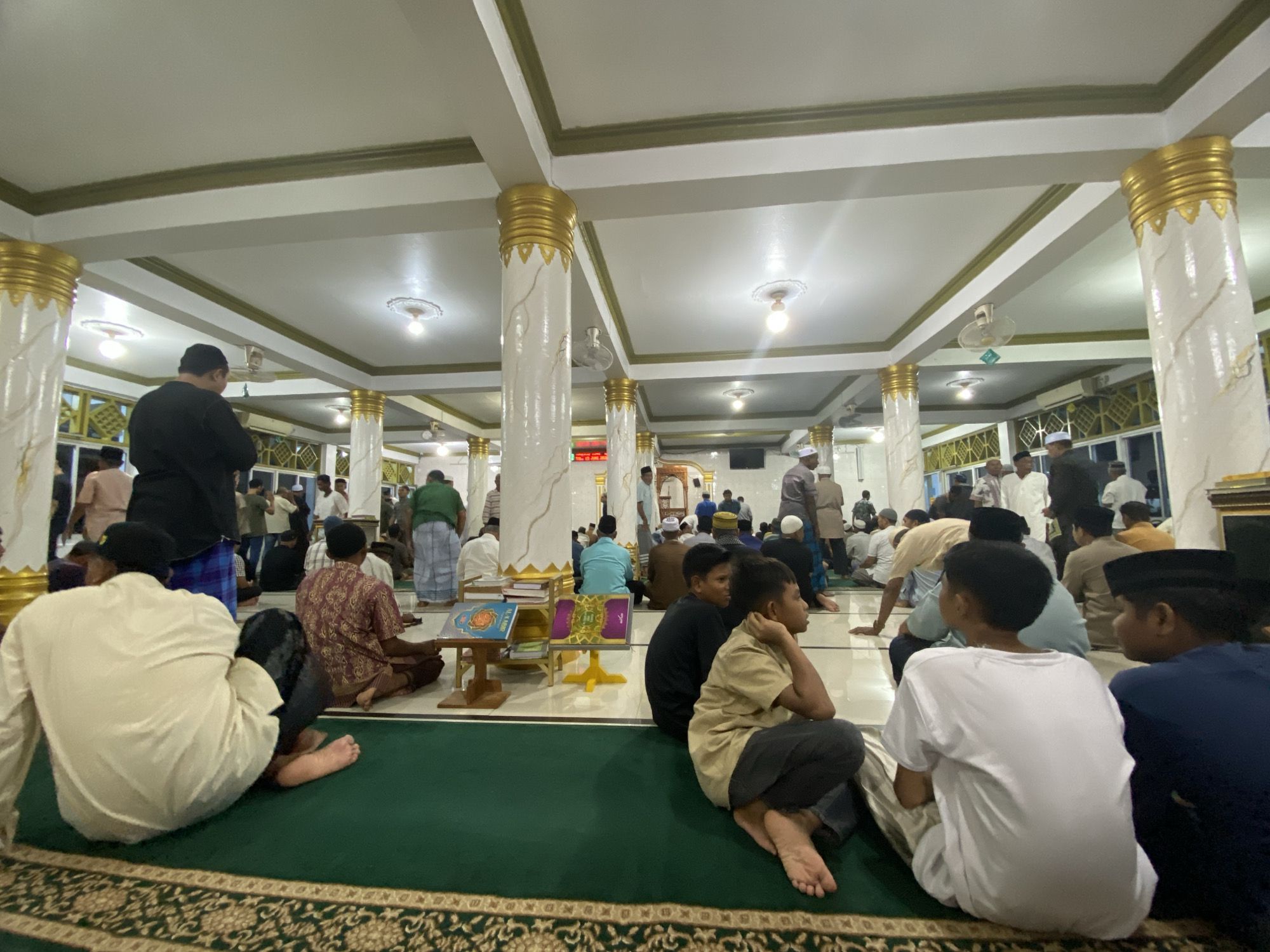 The image shows the interior of a mosque in Lhokseumawe, Indonesia, filled with men and boys dressed in traditional Islamic attire, preparing for prayer. The mosque features a high ceiling with decorative white columns adorned with gold accents, and intricate carpeting in shades of green and gold. Several people are seated on the floor, some reading religious texts, while others appear to be in conversation or waiting. A man in a black robe stands near the entrance, and other individuals are visible in the background, some standing and some seated. The overall atmosphere suggests a communal gathering for religious observance.