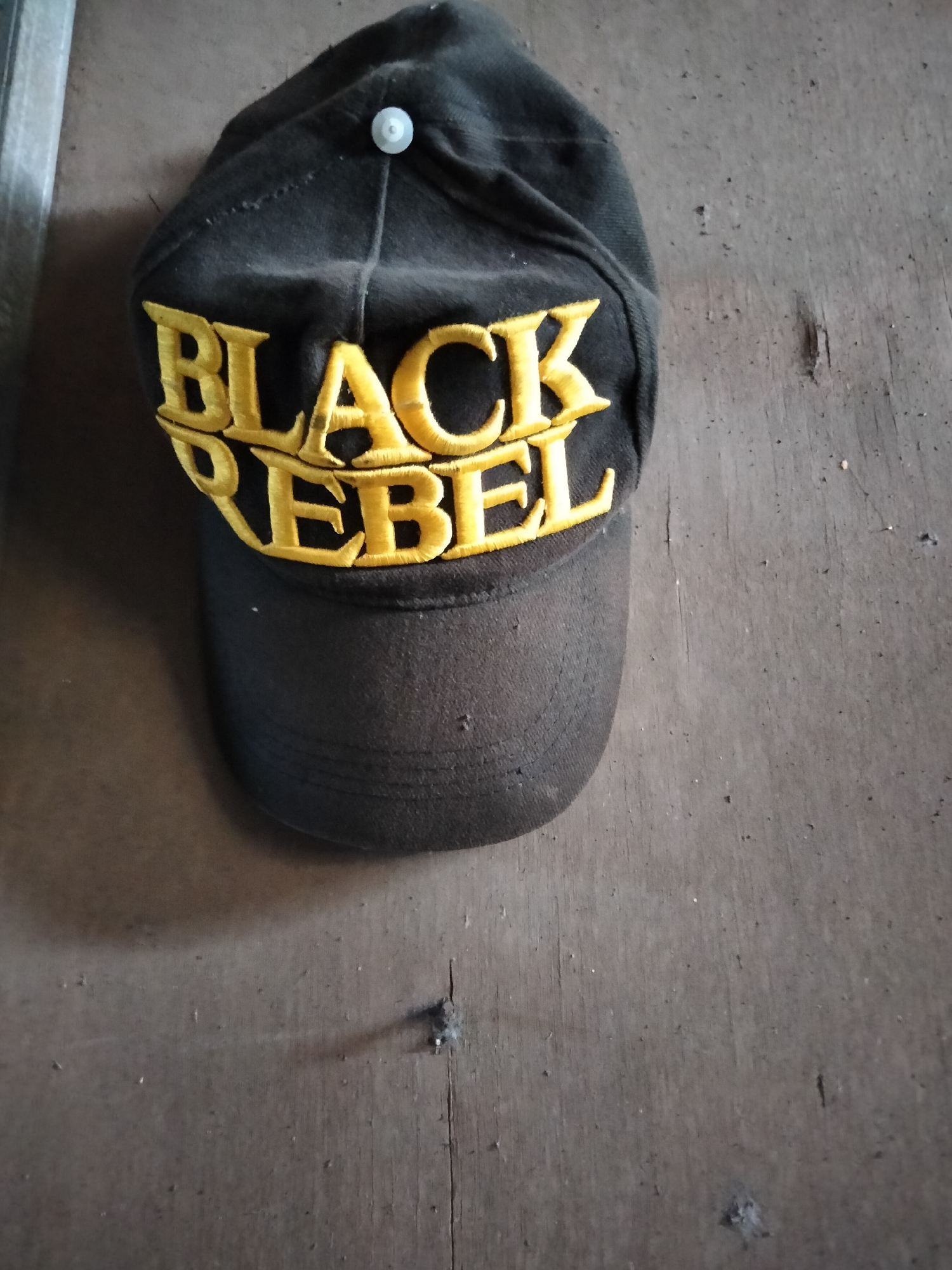 A black baseball cap with yellow embroidered lettering that reads "BLACK REBEL" is hanging on a wooden surface. The cap is positioned at an angle, with the brim facing downwards. The wooden surface appears to be a wall or a fence, with visible grain and several small imperfections. The lighting suggests an outdoor environment, possibly in Denpasar, Indonesia, where such a scene could be found in a residential or commercial area.