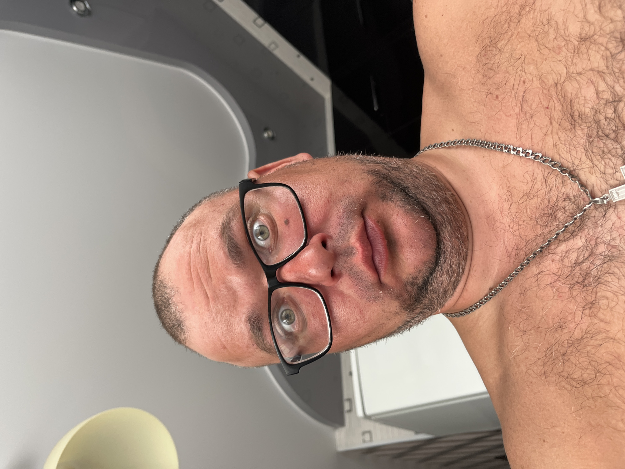 A close-up, eye-level selfie depicts a Caucasian male, estimated to be in his 30s to 50s, looking directly at the camera with a wide-eyed, slightly pursed-lip expression. He is bare-chested, displaying a hairy chest and a silver-colored Cuban link chain necklace with a clear or white Latin cross pendant. The man wears black, rectangular-framed glasses, highlighting his light-colored eyes. He has a balding head with short dark hair visible on the sides and a short, salt-and-pepper beard or stubble. The background reveals an indoor residential setting, with a multi-level ceiling design featuring white, light grey, and darker grey/black sections, some with embedded lighting fixtures. A cream-colored lamp shade is partially visible in the upper left. No specific identifying details are present in the image to determine a location in the city of Minsk, Belarus, or any other specific geographical location.