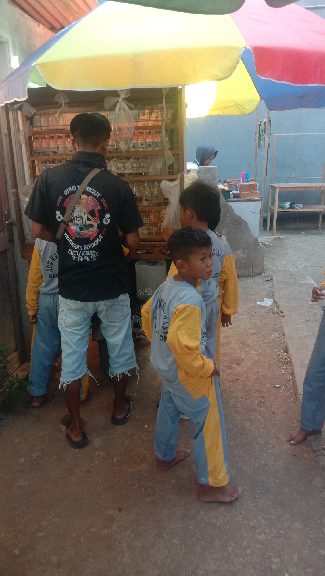 In Karang Anyar, Indonesia, an outdoor scene unfolds beneath a multi-colored umbrella. A man wearing a black t-shirt with text and graphics, and light-blue ripped denim shorts, stands in the foreground. He is facing away from the camera. Three young boys, each wearing a grey shirt with yellow sleeves and light-blue pants, are also present. One boy is directly in front of the man, facing to the right of the frame, while another boy stands to the man's left, facing away. The third boy is partially visible to the right of the frame, also in uniform. Behind the man is a stall displaying numerous small containers, possibly for birds or other small creatures. A woman in a grey hoodie is visible in the background, looking down at something on the stall. The ground is dirt or gravel. Wooden shelves and structures are visible to the right of the stall.