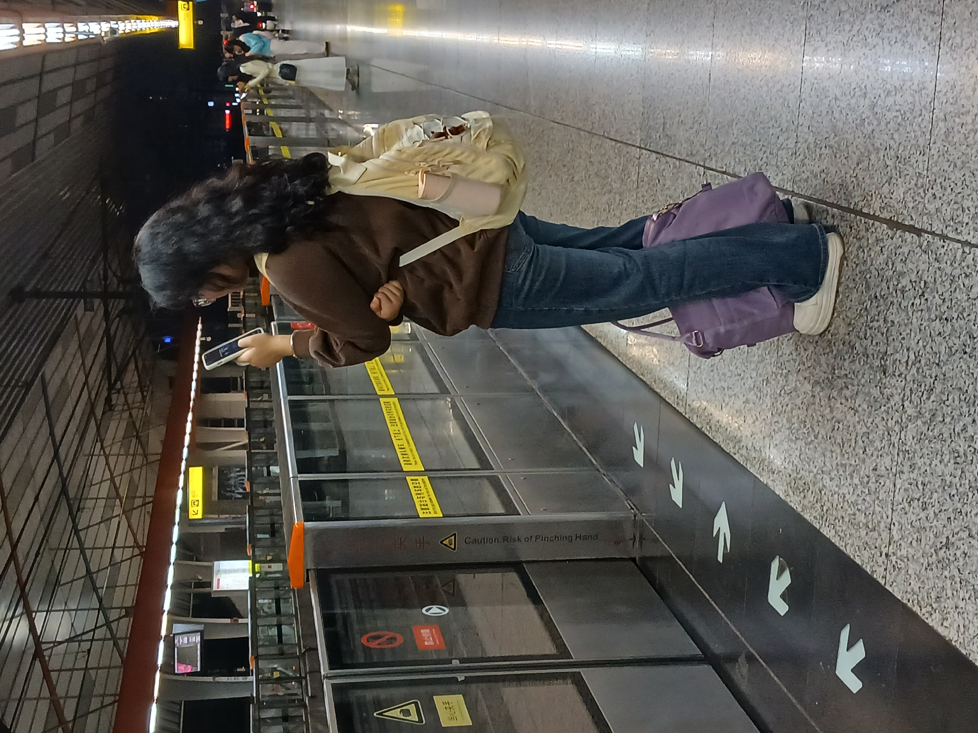 A woman with dark, curly hair stands at a subway platform in the Chongqing High-tech Zone, China, looking at her smartphone. She wears a brown sweater, blue jeans, and white sneakers, and carries a tan backpack and a purple bag. The platform features metal and glass barriers with safety warnings. Arrows are painted on the ground indicating direction. People are visible further down the platform, waiting for a train. The environment is modern, with metallic structures and patterned flooring.