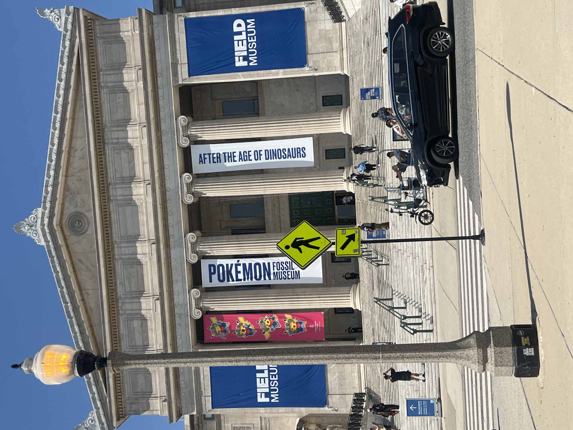 The image shows the exterior of the Field Museum in Chicago. Banners advertising exhibits are displayed on the facade. A black SUV is parked at the top of the staircase. People are walking around. A streetlight and crosswalk sign are in the foreground.