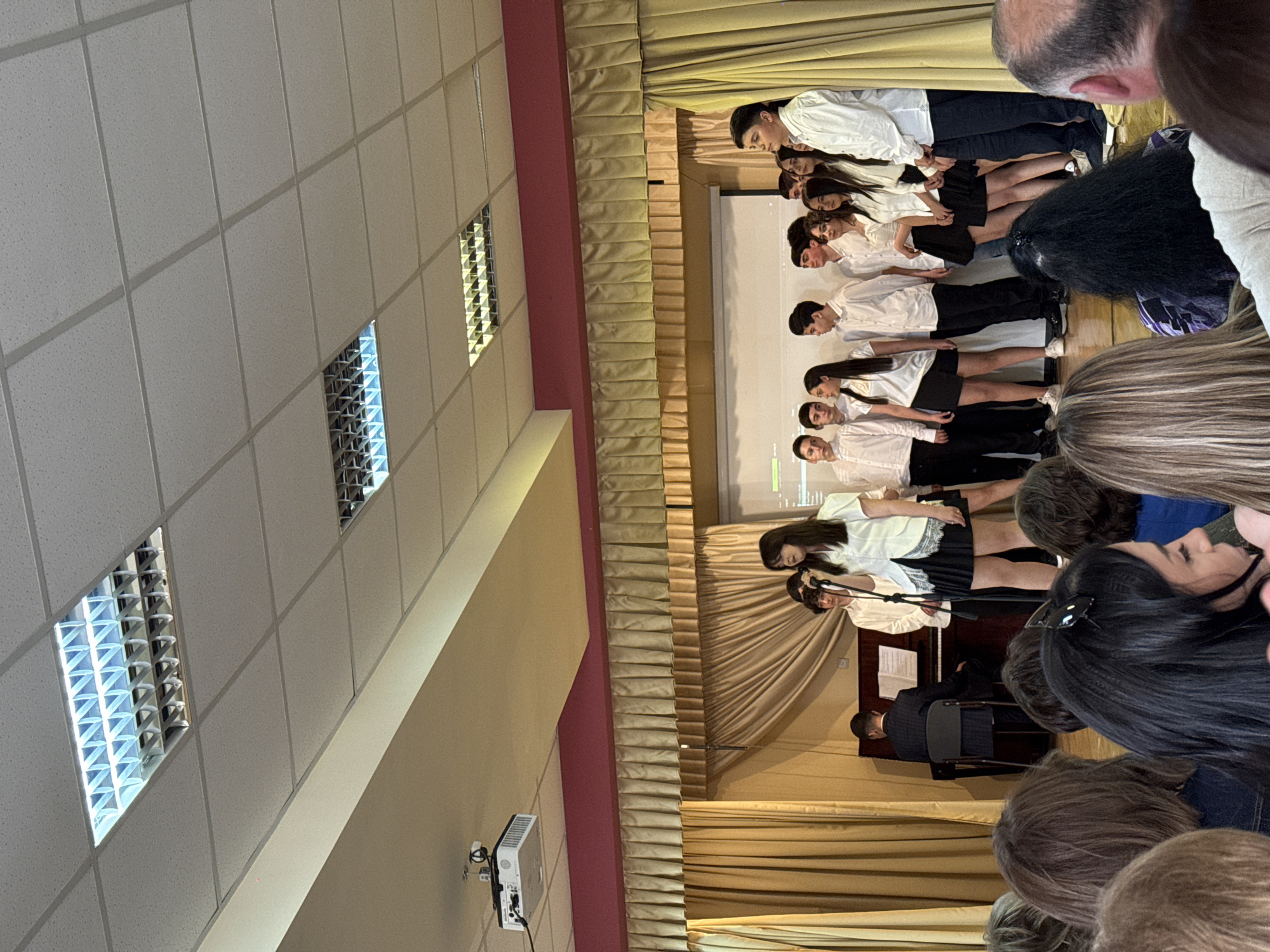 The image shows a group of young people on a stage, possibly a choir or performing ensemble, in Yerevan, Armenia. They are dressed in white shirts and dark skirts or pants, standing in rows. A piano and a pianist are visible at the front of the stage. The audience is partially visible in the foreground, and the background includes stage curtains and a projection screen. The setting appears to be an indoor auditorium or performance hall, judging by the ceiling with recessed lighting.