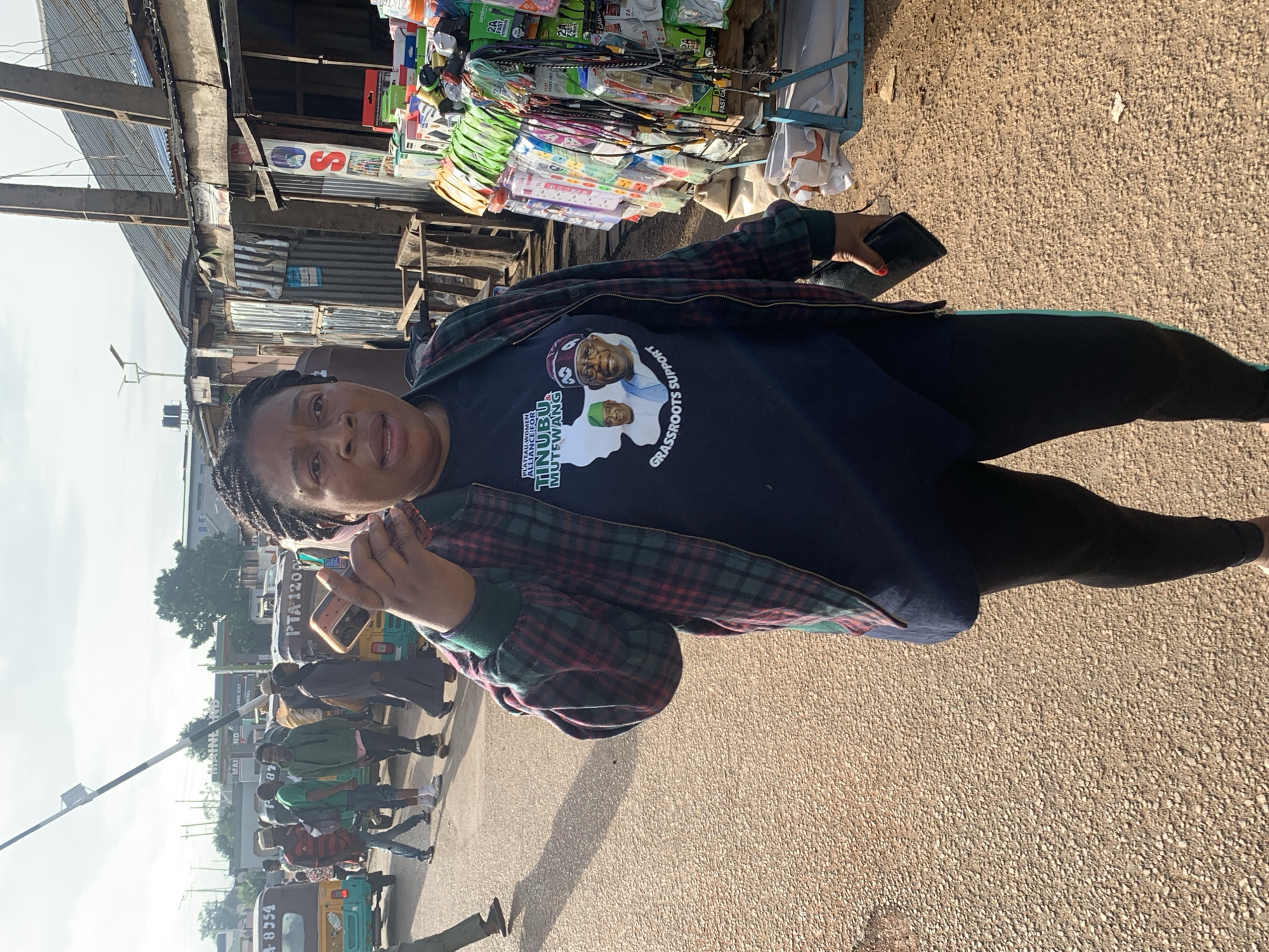 The image captures a street scene in Jos, Nigeria, with a woman in the foreground holding a smartphone to her ear. She is wearing a dark blue t-shirt with a graphic featuring images of two men, a map of Nigeria, and text that appears to read "PLATEAU WOMEN ALLIANCE FOR TINUBU MUTEVANG" and "GRASSROOTS SUPPORT." She is also wearing a plaid bomber jacket and black leggings.
Behind her, the street is busy with activity. Several people are walking, and there are various vehicles, including auto-rickshaws, visible. One auto-rickshaw has "PTA 1208" written on it. On the right side of the image, a market stall is set up, displaying a variety of goods, including electronics like chargers and cables, and bottled drinks. The stall has a corrugated metal roof. The environment suggests an outdoor market area in a bustling urban setting, with paved ground and a mix of commercial structures. The overall lighting indicates it is daytime, with sunlight casting shadows.