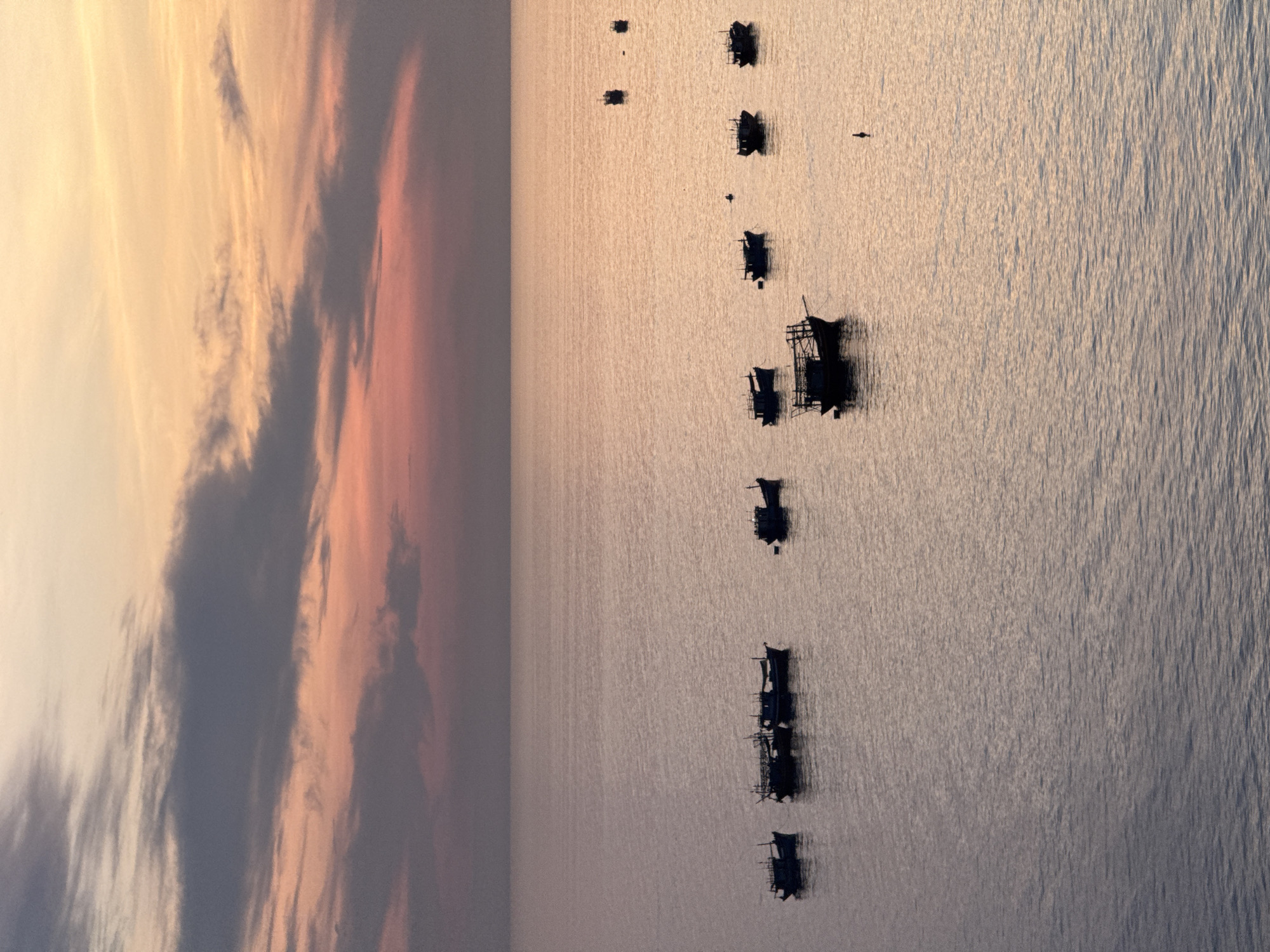 The image depicts a serene seascape at dusk, likely near Lý Sơn Special Administrative Region, Vietnam, given the presence of traditional fishing boats. The sky is a gradient of soft orange and pink hues, transitioning into muted purples and grays, with wispy clouds scattered across the horizon. The calm sea mirrors the colors of the sky, creating a tranquil atmosphere. Several small fishing boats, silhouetted against the water, are anchored or drifting. Some boats are clustered together, while others are more isolated, suggesting a fishing fleet at rest. The overall scene evokes a sense of peace and the quiet end of a day at sea.