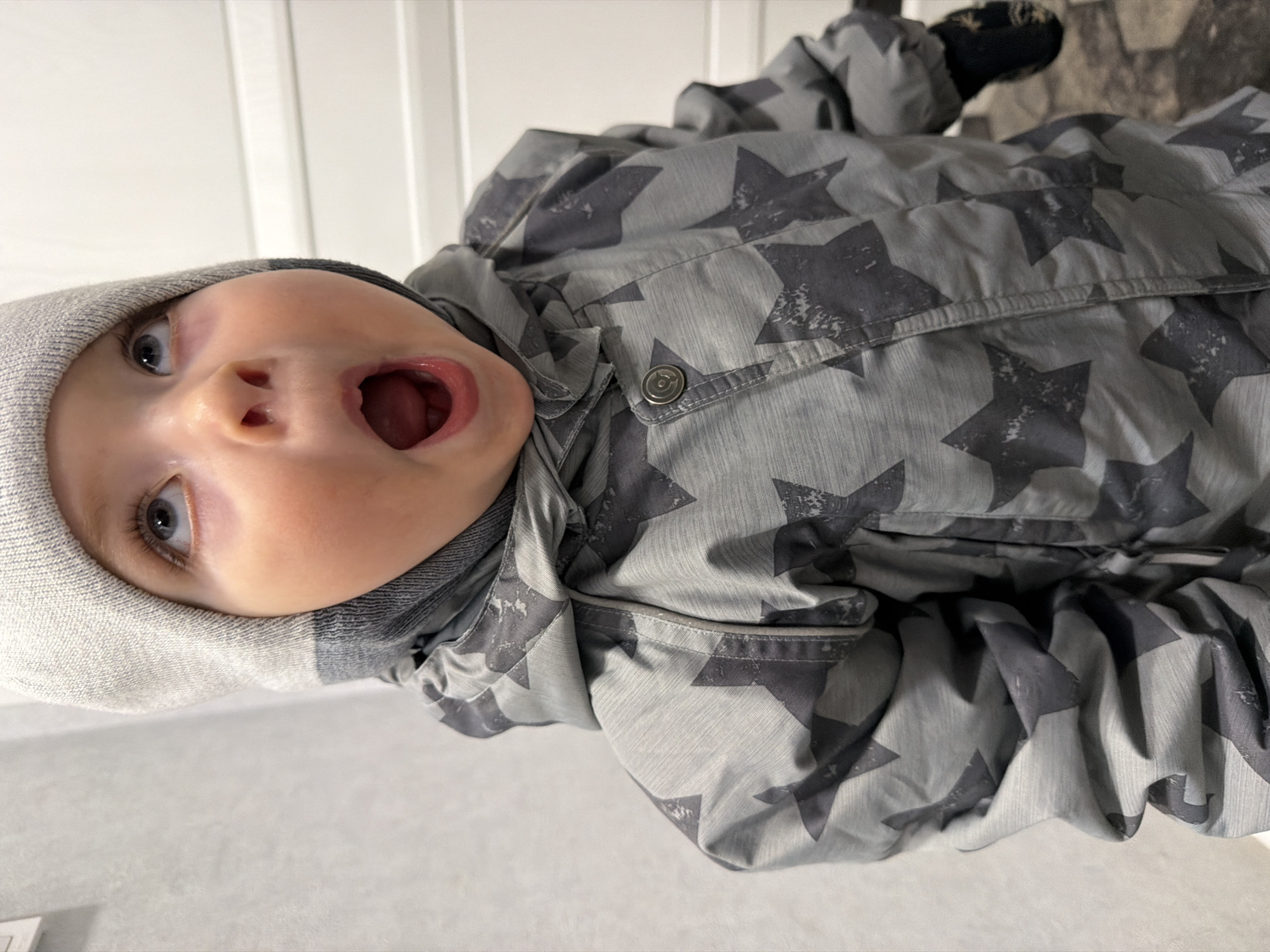 A baby with a gray hat and a gray jacket with a star pattern is lying down with an open mouth. The background is white. It is not possible to ascertain the location as Votkinsk, Russia, from the image.