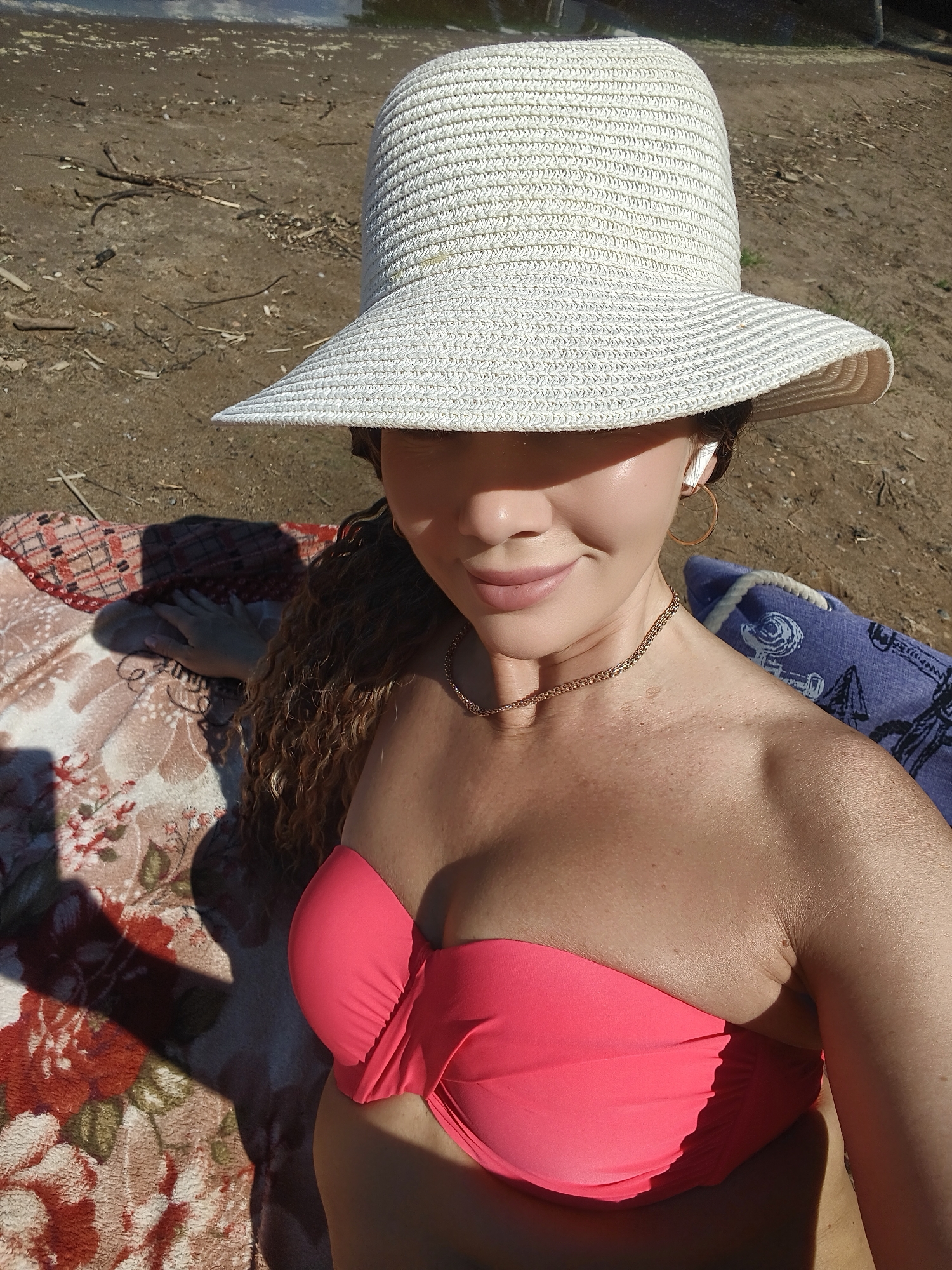 A woman is lying on the beach, wearing a white straw hat, a pink bikini top, and a gold necklace. She has curly brown hair and is wearing hoop earrings and earbuds. She is smiling and looking at the camera. A patterned blanket and a blue towel are behind her. The beach is made of sand and scattered with small branches. The water can be seen in the background.
