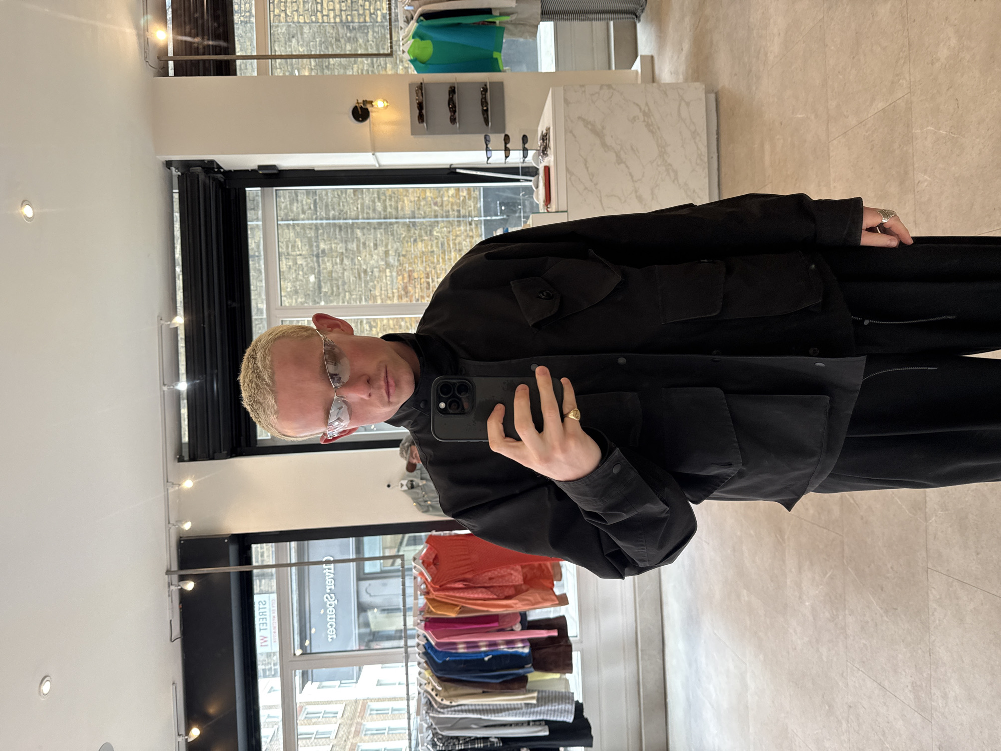 A selfie depicts a fair-skinned individual with short, blond hair and glasses, wearing a black, utility-style jacket, and holding a smartphone with a triple-lens camera. The person also wears a gold ring on their right hand. The scene appears to be within a clothing or eyewear store, evidenced by racks of folded clothing and eyewear displays. Reflected in the window are buildings characteristic of an urban area. A sign in reverse reads "STREET SPENCER MW", suggesting a location on Street Spencer Mews in London, United Kingdom. The floor has a light beige, tiled pattern.