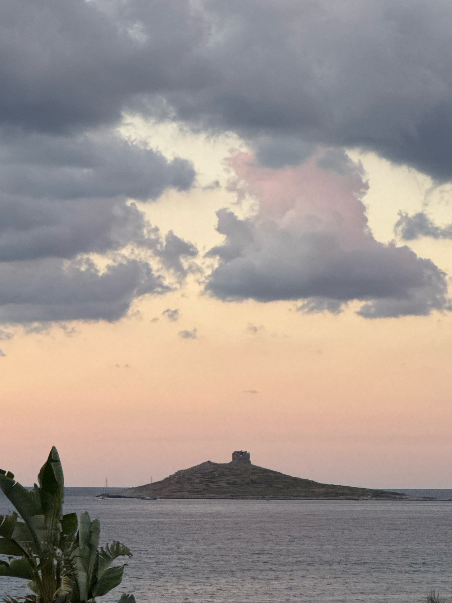 The image depicts a serene coastal scene at sunset in Capaci, Italy. In the foreground, lush green tropical foliage juts into the frame from the left. The middle ground is dominated by a calm, dark blue sea that stretches towards the horizon. On the right side of the middle ground, a small, hilly island rises from the water, topped with the weathered stone ruins of a structure, possibly a watchtower. The background is a gradient of soft, warm colors, transitioning from a pale pink near the horizon to a dusky grey at the top of the frame, indicating the presence of dramatic, textured clouds. Two sailboats are visible in the distance, adding a touch of human presence to the natural landscape. The overall atmosphere is peaceful and picturesque, characteristic of a Mediterranean twilight.