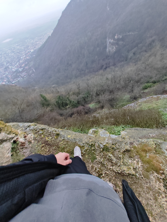 The image is a first-person, downward-angled photograph taken from the edge of a rugged, moss-covered cliff on the Shumen Plateau, overlooking the city of Shumen, Bulgaria.
In the immediate foreground, the lower torso, legs, and feet of a person are visible. The individual wears black pants, a grey upper garment, and a dark (likely black) jacket, with light grey or white sneakers on their feet. Their right hand rests casually on their right thigh.
Directly below the person's feet, a steep, heavily wooded slope descends into a valley. The trees are predominantly deciduous and bare, indicating a colder season, with patches of green undergrowth and dry brush. Further in the background, to the lower left, a sprawling urban area, identified as the city of Shumen, is visible with numerous buildings featuring reddish roofs, partially obscured by atmospheric haze. A large, densely forested mountain or hillside occupies the upper right background, merging into an overcast, hazy sky. The overall environment is one of a high, natural overlook above a cityscape.