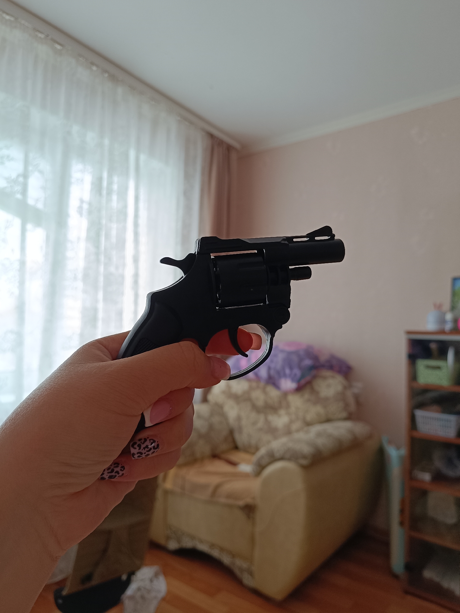 A right hand holds a black revolver-style handgun, with the index finger near the trigger, indoors. The hand has fingernails painted with leopard spots and a white French tip. The background features a beige armchair with floral accents, a wooden floor, a partly visible white curtain and a cabinet with various items. The location in the city of Tobolsk, Russia, is not discernable from the information provided in the image.