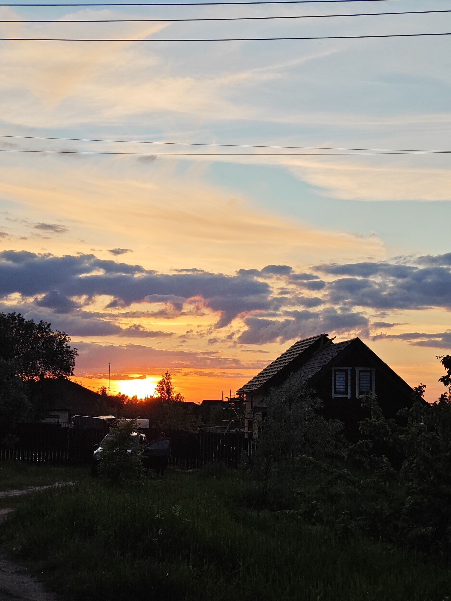 The image depicts a serene sunset scene in Rybolovo, Russia, characterized by a dramatic sky and silhouetted rural features. The upper part of the sky shows light blue hues with wispy, pale orange clouds, while the mid-section transitions into darker grey clouds interspersed with vivid oranges and yellows from the setting sun. Several thin, dark power lines stretch horizontally across the upper sky.
The sun, a brilliant orange sphere, is partially visible on the horizon towards the left, nestled behind silhouetted trees and distant structures. In the foreground and midground, a dark silhouette of a village or rural area is visible. On the right, a house with a dark, sloped roof and two illuminated rectangular windows on its upper floor stands prominently, possibly with scaffolding or construction elements on its left side. To the left, a dark wooden picket fence extends horizontally, behind which a dark-colored car with an open door is partially seen amidst dark bushes. Another partially visible, silhouetted house or structure is further to the left.
The lower foreground features green grass and a dirt or gravel path in the bottom-left corner. Numerous trees and bushes, rendered in dark silhouette, frame the scene and the setting sun. The time of day is clearly sunset, creating a tranquil yet visually striking atmosphere. No people or visible text are present in the image.