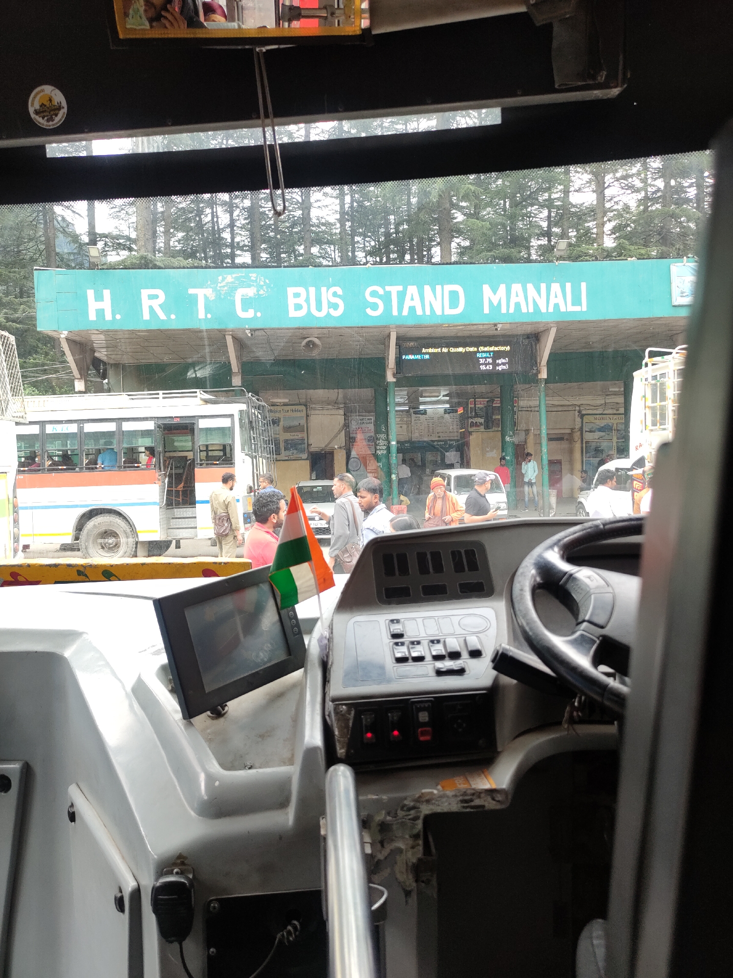The image is taken from inside a bus, looking out through the windshield at the HRTC Bus Stand in Manali, India. A green sign with white lettering reads "H. R. T. C. BUS STAND MANALI." Below the sign is a digital display showing "Ambient Air Quality Data (Satisfactory)." Several people are visible outside the bus, including a man in a light brown uniform, a man in a pink shirt, and others. A bus with "HRTC" written on it is parked at the stand, with its door open and people boarding or disembarking. An Indian flag is visible in the foreground, placed on the dashboard. The environment appears to be a mountainous region, with pine trees visible in the background. The interior of the bus shows a dashboard with various buttons and a steering wheel.