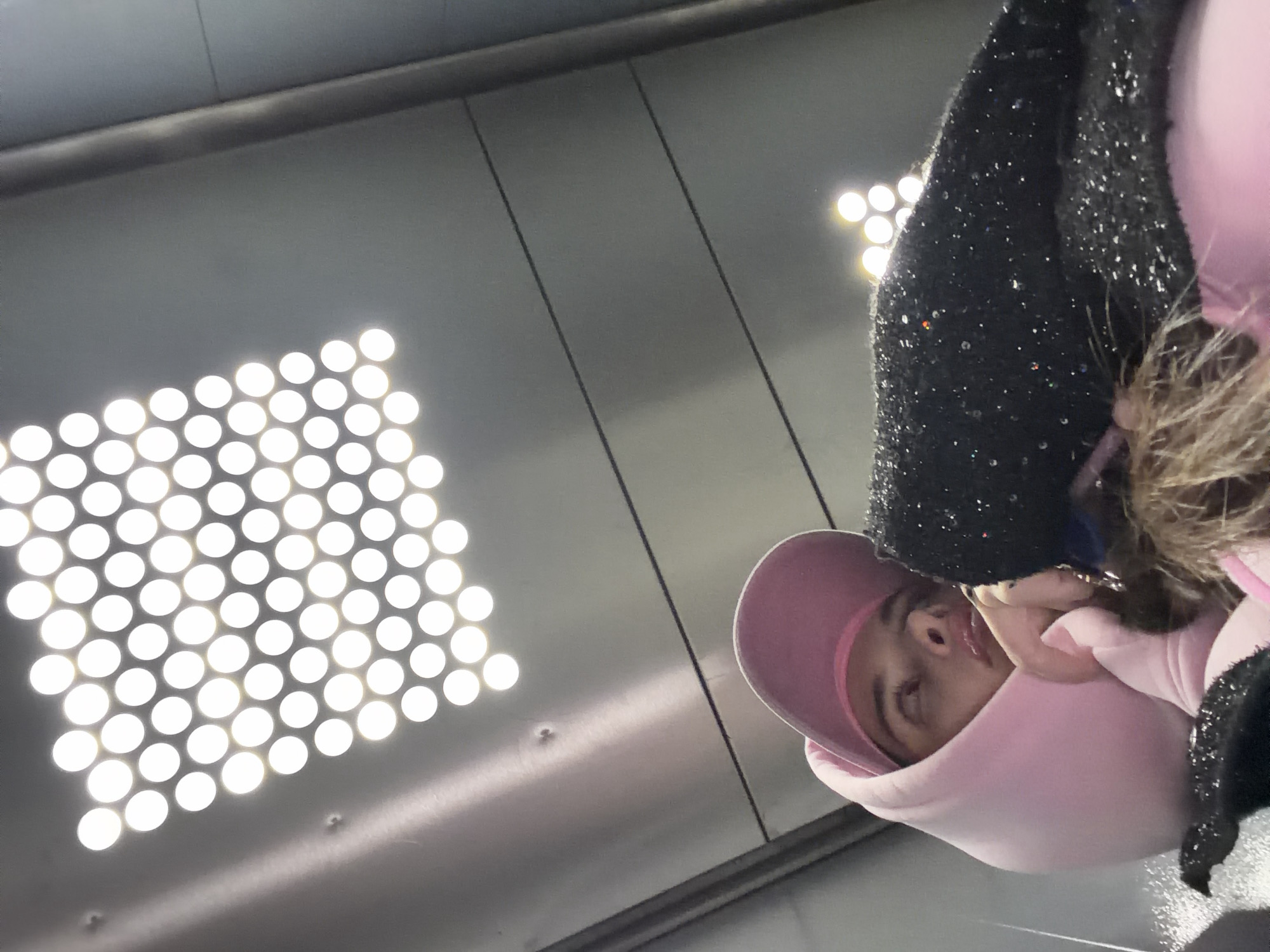 A person with light skin is visible, wearing a pink hoodie with the hood up and a black sparkly coat, positioned in the lower right quadrant of the image. The camera angle is from below, looking upwards towards the subject. The environment is a grey, industrial-looking space with a grid of evenly spaced, round lights in the upper left quadrant, suggesting a modern or underground setting. The location is not evident from the image.