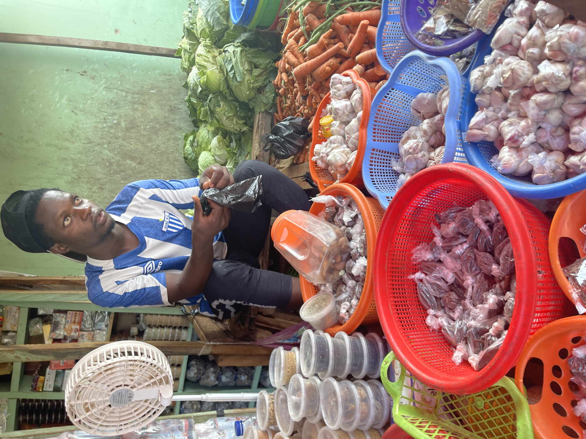 A man wearing a blue and white striped soccer jersey and a black cap sits in a market in Akwanga, Nigeria. He is bagging something from a basket filled with various small packages wrapped in clear plastic. The scene is filled with an assortment of produce and packaged goods.
To the left of the man are heads of cabbage stacked high. Behind him, carrots are piled up, interspersed with baskets of garlic. In the foreground, there are baskets containing more small packages similar to the ones he is bagging, as well as containers of spices. A small white fan hangs in the lower left corner, and shelves of goods are visible behind him.