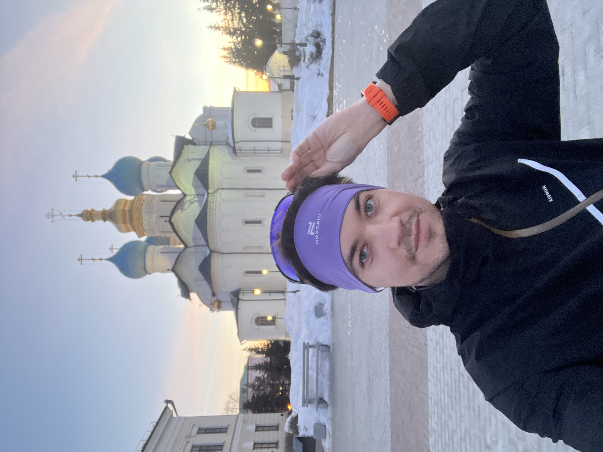 A male individual is depicted in a selfie-style shot in front of the Annunciation Cathedral (Blagoveshchensky Sobor) within the Kazan Kremlin, Kazan, Russia. The man, appearing to be in his 20s or 30s, has light-colored eyes and stubble, and is looking directly at the camera with a slight smile. He wears a black outdoor jacket with "KIPRUN" visible on the chest and white stripe detailing. A purple headband with the "NORDSKI" logo and dark sports sunglasses are worn pushed up onto his head. An orange smartwatch is on his left wrist, and his left hand is raised, adjusting his headband or hair.
The background features the white-walled Annunciation Cathedral with its prominent central golden onion dome and two flanking blue onion domes, all topped with Orthodox crosses. The scene is set during winter, indicated by significant snow accumulation on the ground, piled high in drifts. The sky displays light blue at the zenith, transitioning to warm orange and pink hues near the horizon, suggesting sunset or twilight. Artificial streetlights with warm illumination are visible among snow-dusted coniferous trees to the right of the cathedral. A paved ground surface, partially covered in snow or ice, occupies the immediate foreground. A grey park bench is partially visible, partially buried in a snowdrift, to the left of the central background.