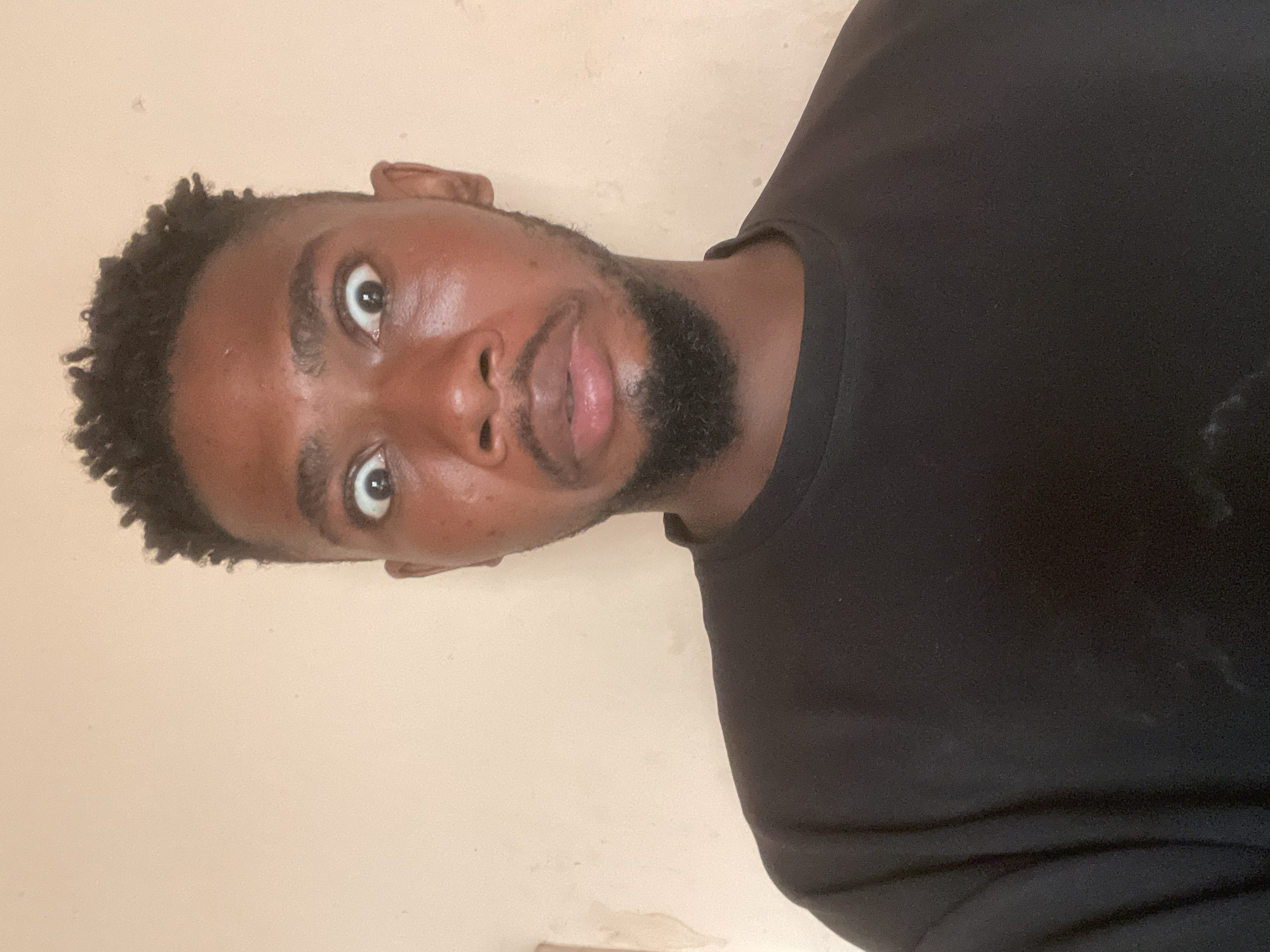 A medium-close-up headshot photograph shows a man of African descent with short, dark, tightly coiled hair, a dark beard, and dark eyes looking slightly upward with a surprised expression. He has smooth, dark skin and is wearing a black t-shirt. He is positioned slightly to the left. The background is a plain, light beige wall. This image was not taken in Unguwar Gabas, Nigeria.