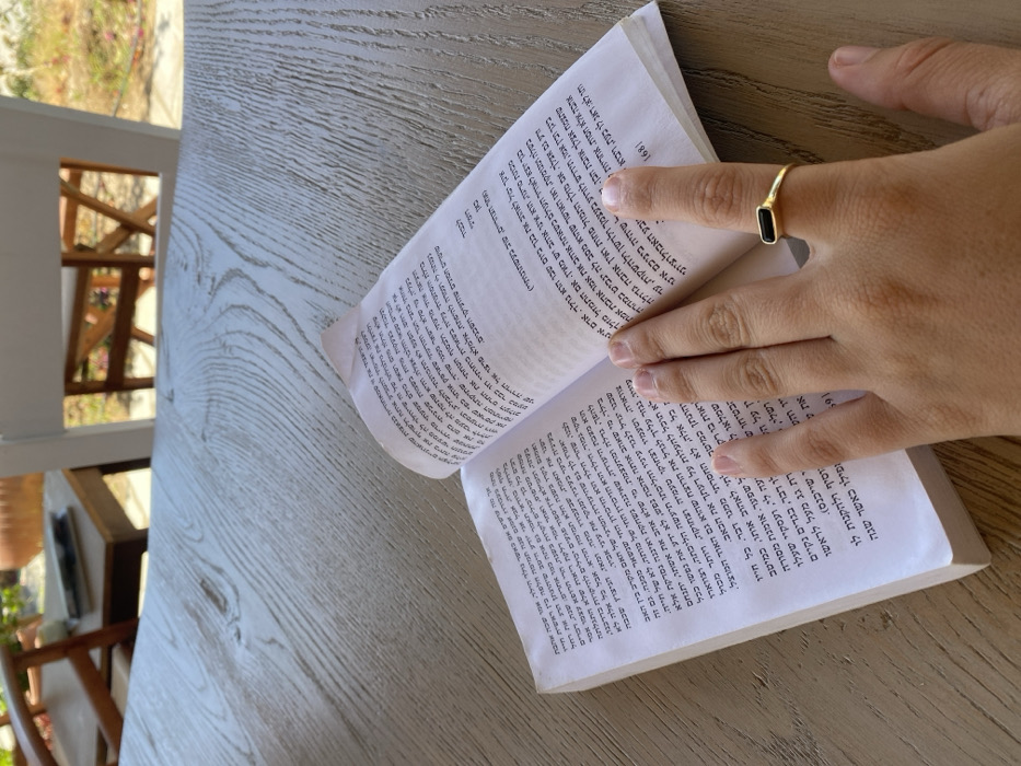 An overhead photograph captures an open book resting on a light-colored, weathered wood-grain table. A human hand, positioned with fingers splayed, rests on the lower right page of the book. The hand wears a gold-toned ring with a black, rectangular, horizontally set stone on the ring finger. The book's pages display text in Hebrew script, with the page number "168" visible at the bottom right corner of the right-hand page. The background is out of focus, revealing elements consistent with an outdoor or semi-outdoor setting, including parts of a white wooden chair, a natural wood-colored chair, green foliage, and a terracotta pot, indicating a well-lit environment. The scene is located in Πλατύς Γιαλός, Greece.