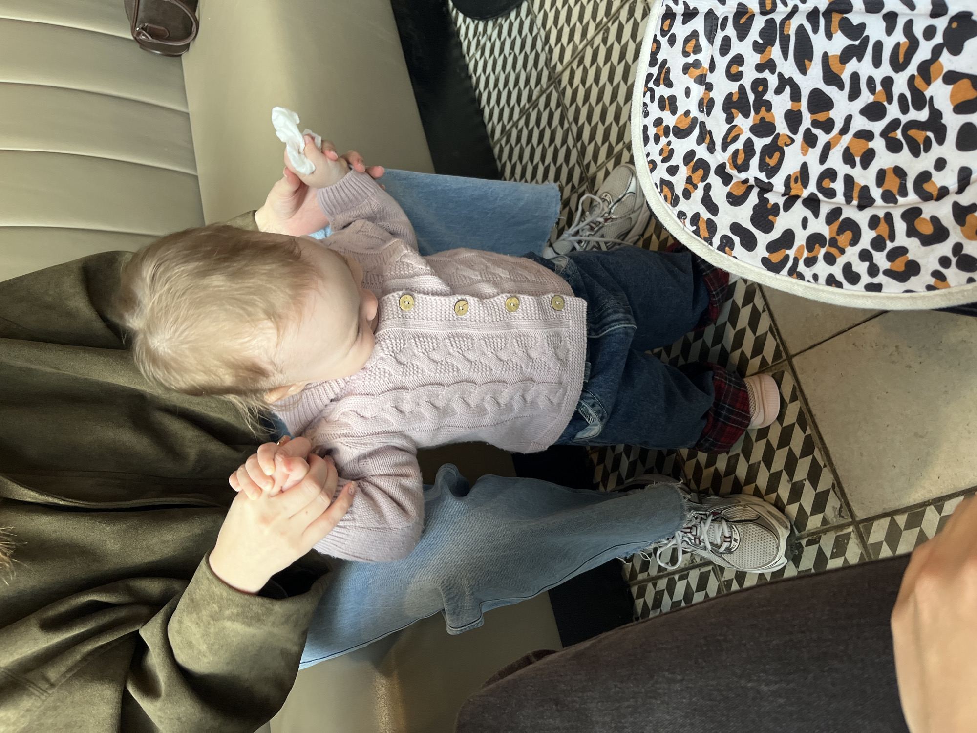 A baby wearing a light purple sweater and jeans sits in the lap of a person wearing a green jacket and blue jeans. The baby is holding a white cloth in its hand and the adult is holding the baby's other hand. The scene is indoors. There is a table with a leopard print cover and sneakers on the floor. The floor tiles are black and white. The location of Shushary, Russia, is not discernible from the image.