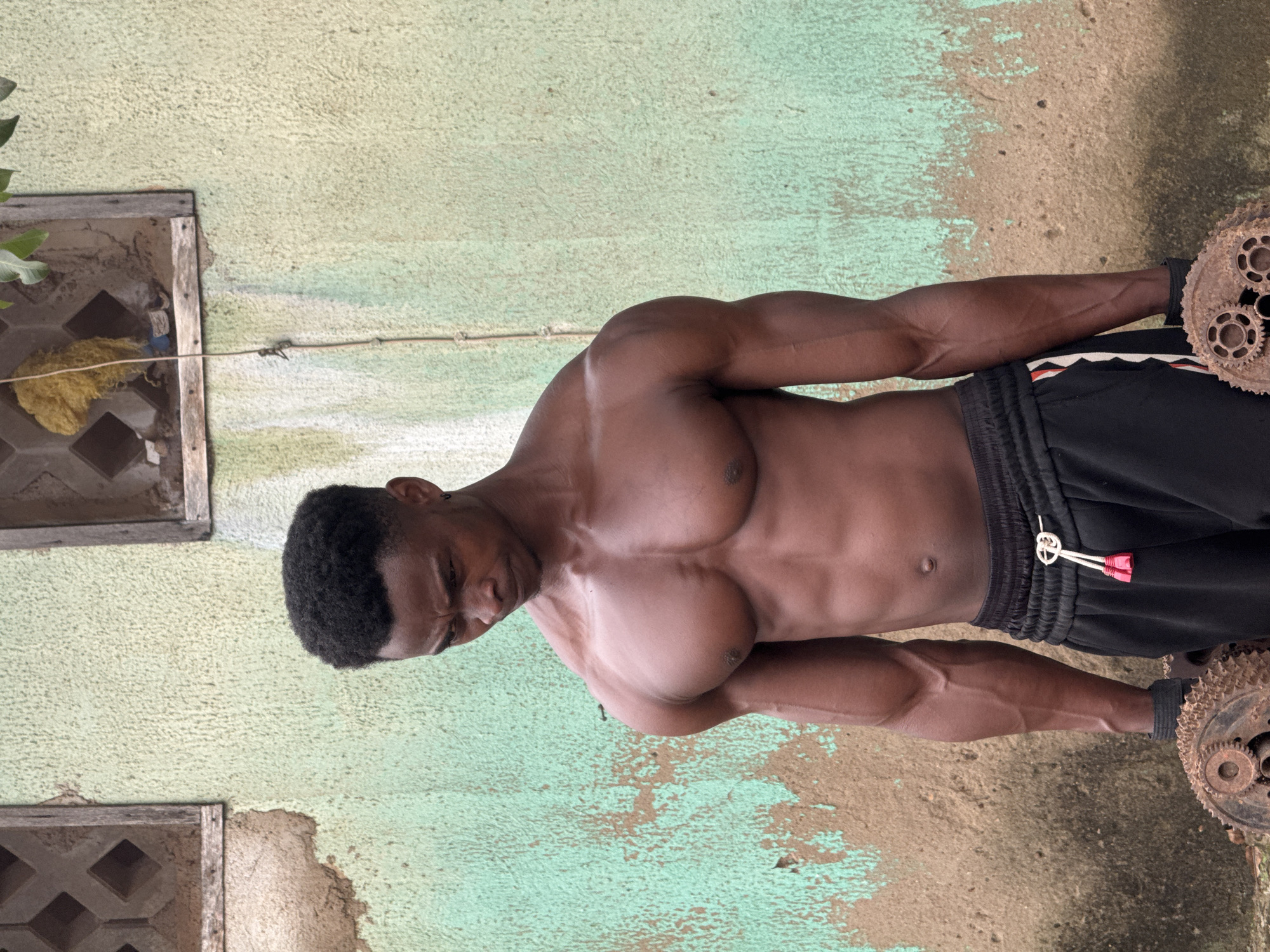 A shirtless, muscular Black man stands outdoors in front of a textured, weathered wall that is painted a faded green. The man is looking down and to his right, with his left arm bent and holding a rusty, gear-like object. His right arm is also bent and holding a similar object. He is wearing black athletic shorts with a white drawstring. The wall has two rectangular openings at the top, resembling windows, with wooden frames. One opening contains a bundle of yellow material. The ground appears to be dirt. The scene suggests a training or workout session in a somewhat rustic outdoor environment, possibly within Akwanga, Nigeria, given the architectural and environmental clues.