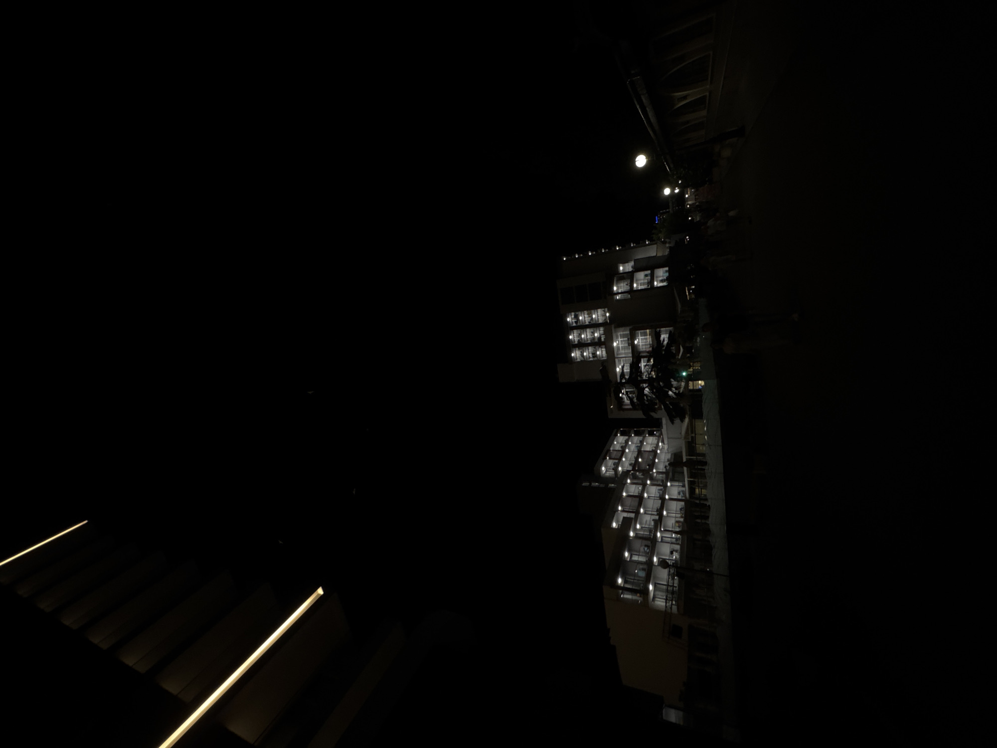 The image captures a nighttime scene in Varna, Bulgaria, likely outdoors. A modern, multi-story building with many lit windows dominates the right side of the frame. The windows appear to be on balconies or within rooms, suggesting it might be a residential building or a hotel. Lights from inside the building create a warm glow, contrasting with the dark night sky.
Above the building, a bright, circular object is visible, which could be the moon or a strong streetlamp. A series of streetlights are also discernible, arranged in a line, further illuminating the area.
On the left side of the image, there are angled structures with illuminated strips, possibly part of a modern architectural feature or an outdoor seating area. The overall mood is one of quiet urban night, with the artificial lights of the building and street fixtures cutting through the darkness. The image is oriented sideways, with the building appearing to run vertically. No people are clearly visible, but the lit windows imply activity within the building.