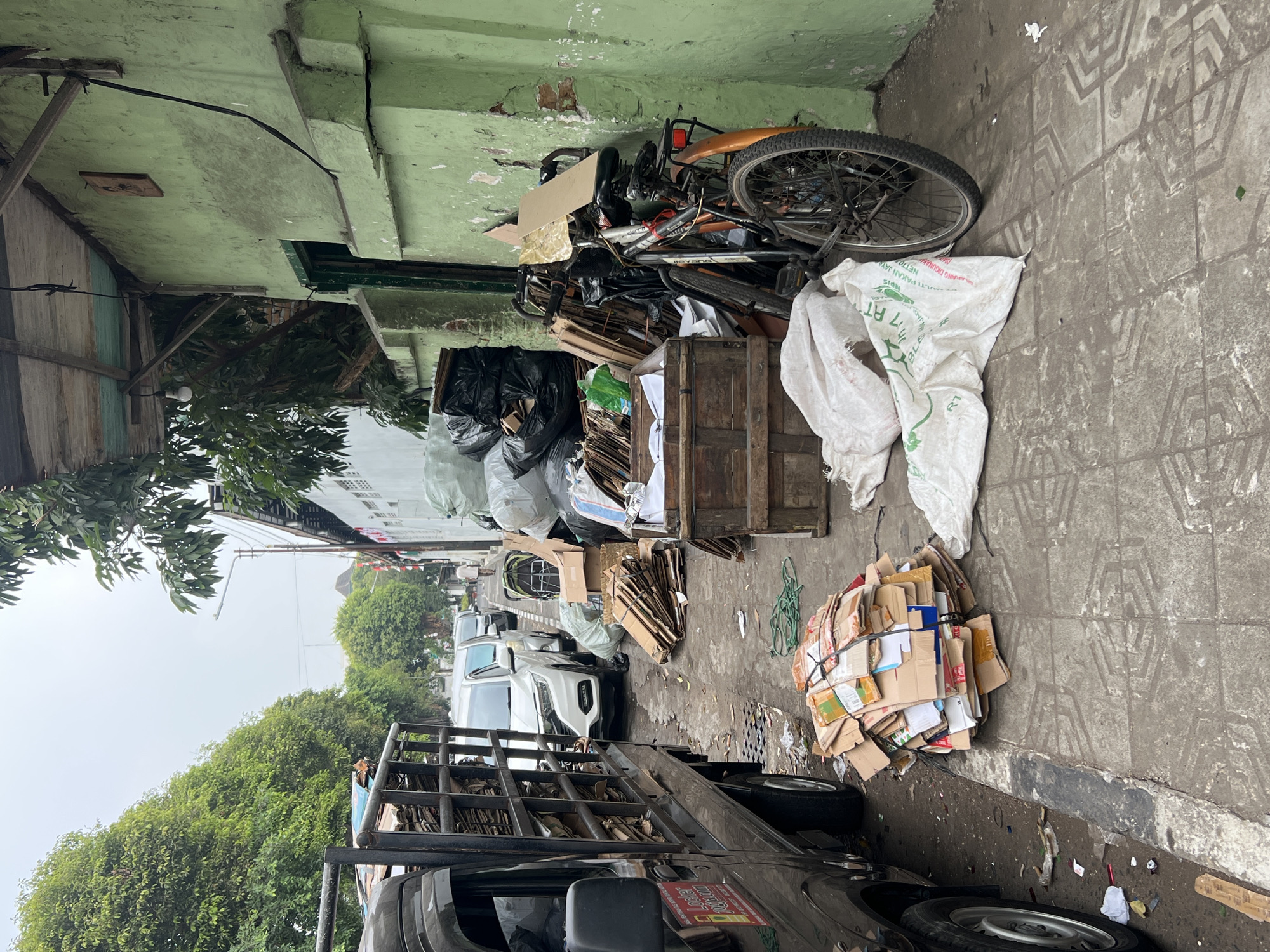The image depicts a street scene, likely in Yogyakarta, Indonesia, characterized by a significant amount of collected recyclable materials. A black utility truck is parked on the left, loaded with stacks of flattened cardboard boxes. In front of the truck, a pile of similarly bundled cardboard is placed on the sidewalk.
The sidewalk is lined with various items: several large white sacks, some appearing to contain more recyclables, are spread out. A wooden crate filled with paper and cardboard is situated near the center of the frame. Behind the crate, a bicycle with an orange fender and basket leans against a weathered, light green building. The building itself appears aged, with peeling paint and a visibly old wooden overhang above the doorway.
Further down the street, other vehicles, including a white SUV and a silver sedan, are visible, suggesting it's a passable street, possibly in a residential or commercial area. The overall environment is cluttered, with scattered debris on the sidewalk and a general sense of disarray, consistent with a collection point for recycled goods. The sky is overcast, indicating a cloudy day.