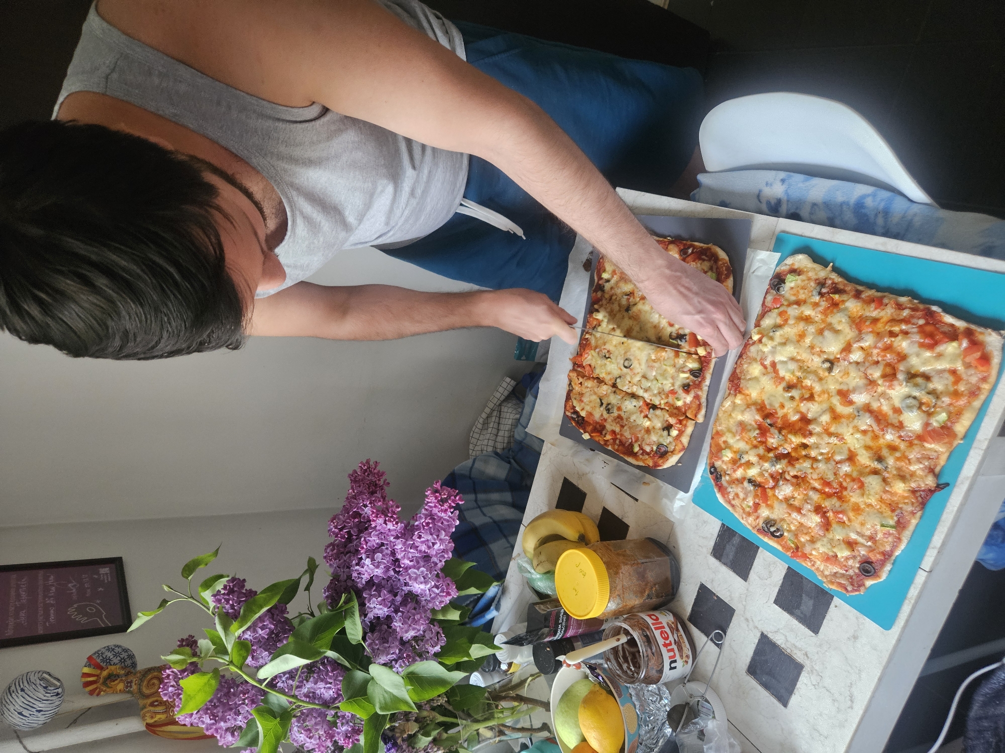 A person is cutting a rectangular pizza into slices in a kitchen setting. Two pizzas are visible: one on a gray surface and the other on a turquoise cutting board. The person is wearing a gray tank top and blue pants. A bouquet of purple lilacs sits on the counter near a collection of items including bananas, a jar with a yellow lid, a jar of Nutella, and various fruits in a bowl. The kitchen has a light-colored countertop with black squares, and there is a white chair partially visible. The location is potentially Rodniki, Russia, as indicated by external information.