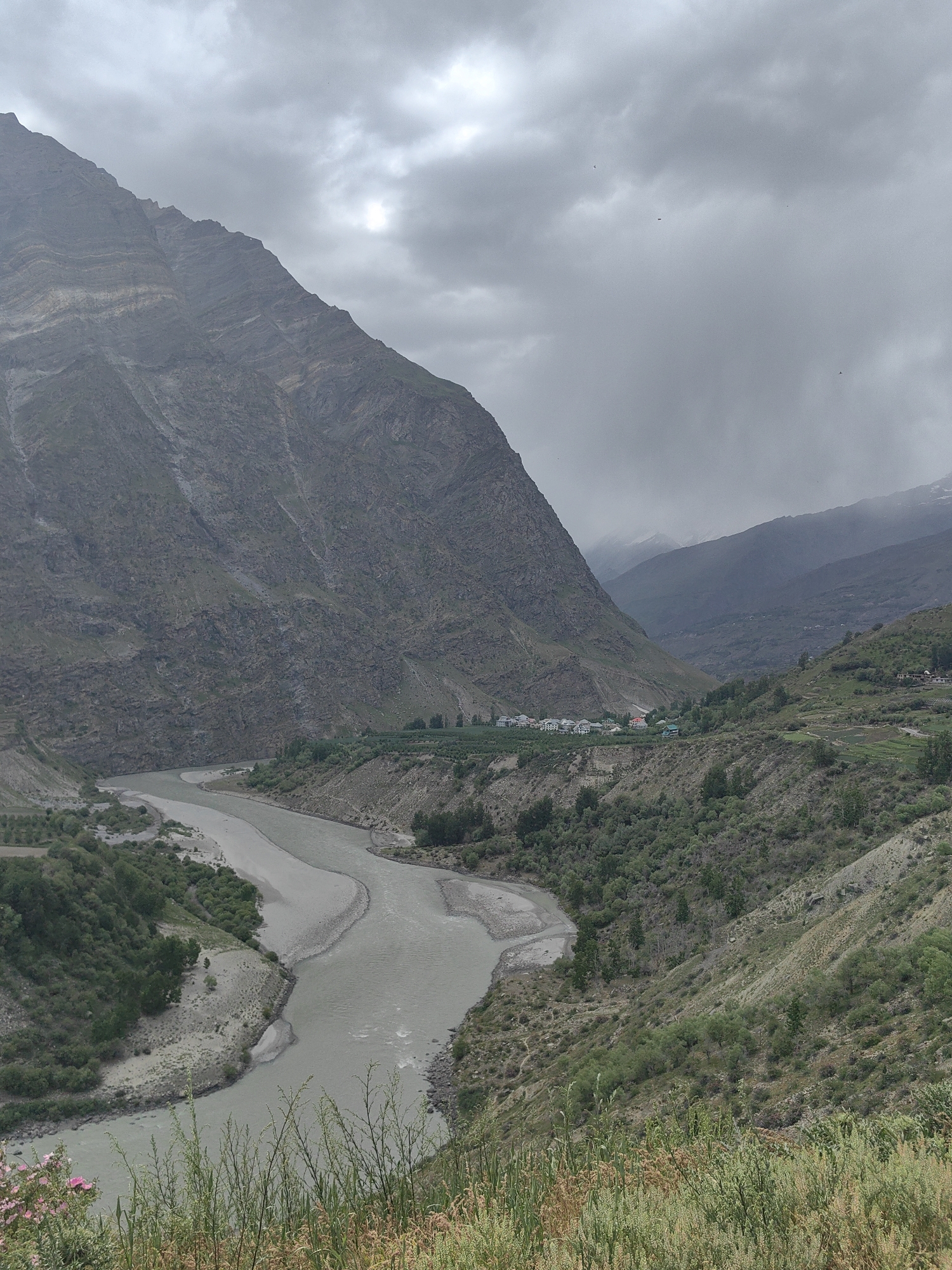 The image depicts a wide river flowing through a valley in Tandi, India. The river, appearing murky, curves through the landscape, bordered by steep, rocky mountains on the left and gentler, vegetated slopes on the right. The mountains exhibit distinct geological layers. A small village with white-roofed buildings is nestled in the valley on the right side, surrounded by greenery and agricultural fields. The sky is overcast with dramatic, grey clouds, suggesting a cloudy or possibly rainy day. In the foreground, there is lush green vegetation and some pink flowers. The overall scene conveys a sense of natural grandeur and rural tranquility.