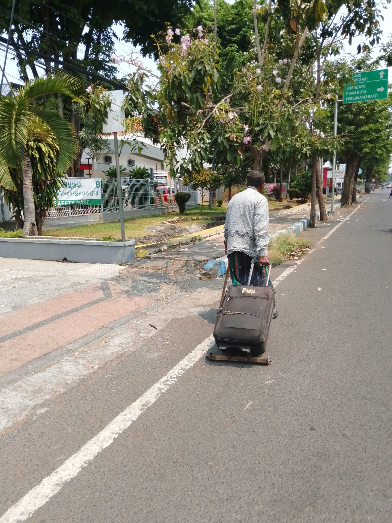 A man wearing a white patterned long-sleeved shirt, a dark cap, and a sarong walks along the side of a paved road in Pasuruan, Indonesia, pulling a large dark suitcase. The suitcase has been modified with a wooden plank and a wheel attached to its base to facilitate dragging. The man is walking on the asphalt, adjacent to a sidewalk with a concrete curb and a narrow strip of grass and dirt. Beyond the sidewalk, there is a fence and buildings, including one with a banner displaying text and a safety logo. Trees, some with pink flowers, line the area. A green directional sign is visible in the background, indicating directions to "WOSARI," "SURABAYA," "PDAM KOTA," and "PERUMKARYA BAKTI." The scene is outdoors, under daylight conditions, with shadows indicating a sunny day.