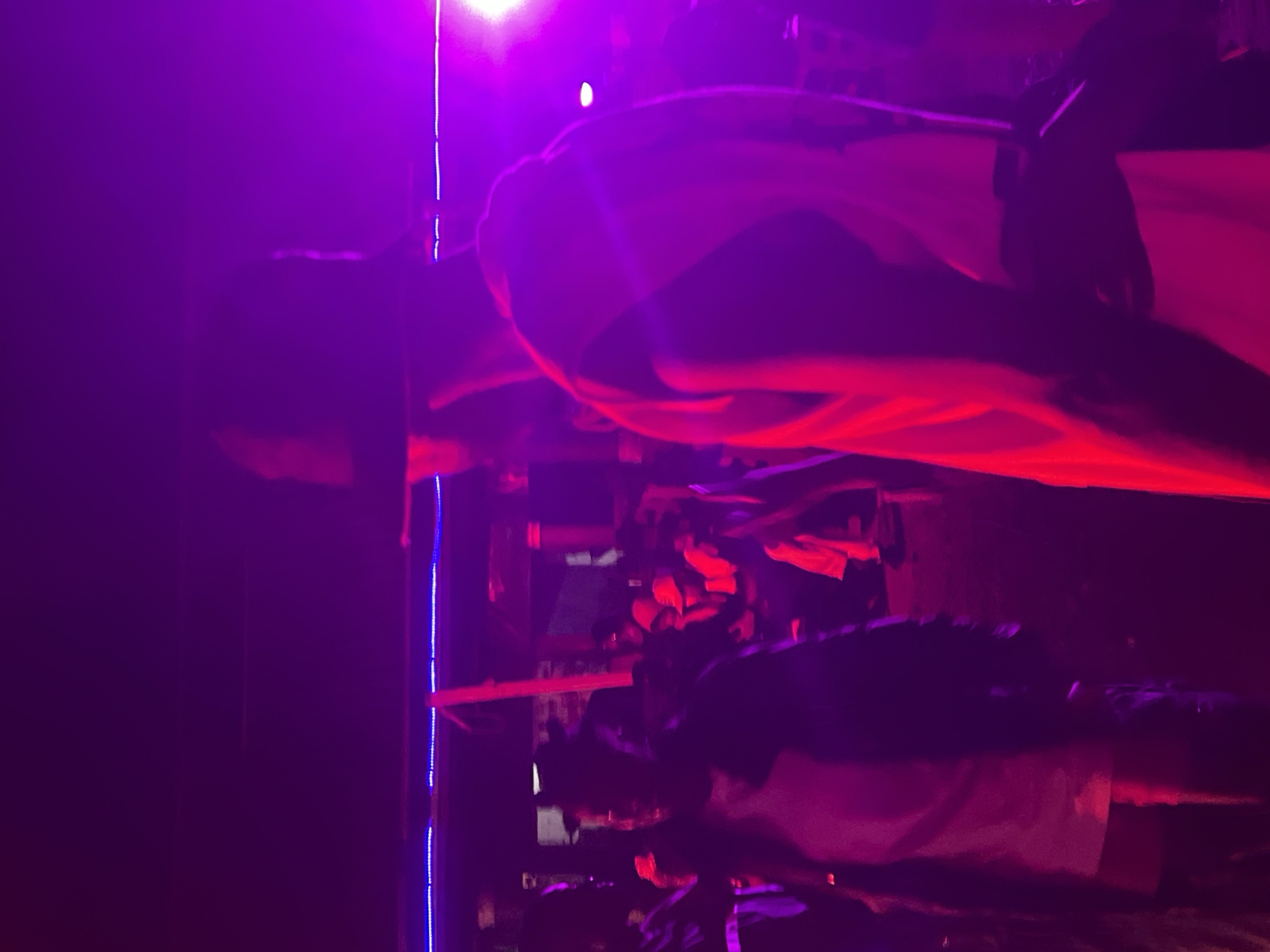 The image shows a dimly lit outdoor gathering at night in Durumi, Nigeria, illuminated by vibrant purple and red stage lights. Several individuals are present, some of whom are blurred due to motion or low light conditions. In the foreground, a person wearing a hat and a light-colored garment with dark trim is the most prominent subject. Their body is angled away from the camera, with their right arm visible. In the mid-ground, another individual, also partially obscured, appears to be wearing a dark outer garment over a lighter shirt. Further in the background, a crowd of people are gathered, with some wearing hooded tops in orange and white. The environment suggests a festive or performance setting, possibly an outdoor concert or street event, with elements of structure and lighting visible, including what appears to be a tent or awning overhead and decorative blue lights. The overall atmosphere is energetic and social.