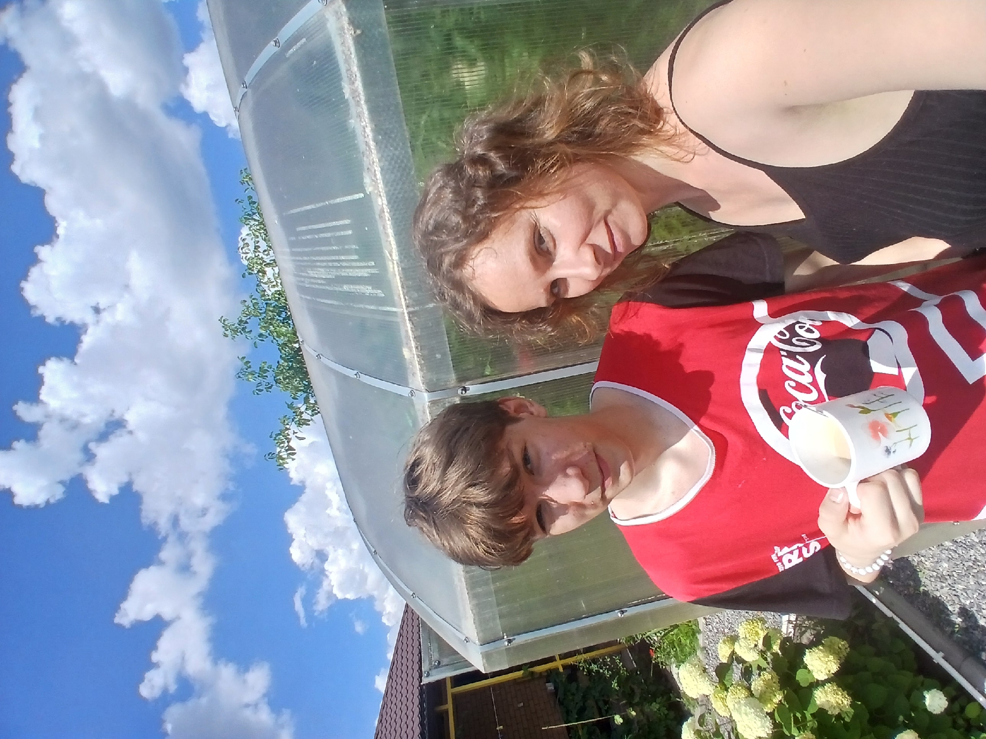 A medium shot captures a young man holding a mug, and a woman standing next to him, both positioned in front of a greenhouse. The young man, on the left, is wearing a red t-shirt with a Coca-Cola logo and a beaded bracelet on his left wrist. He is holding a white mug decorated with floral patterns. The woman, on the right, is wearing a black tank top. They are outdoors with a bright blue sky filled with fluffy white clouds above. A greenhouse with a translucent roof is behind them, and to the left, a brick house and lush green foliage are visible. The ground is covered with gravel. The scene appears to be a residential area, possibly a dacha or a house in Maslova Pristan', Russia, given the presence of a greenhouse and garden elements. The lighting suggests a sunny day.
