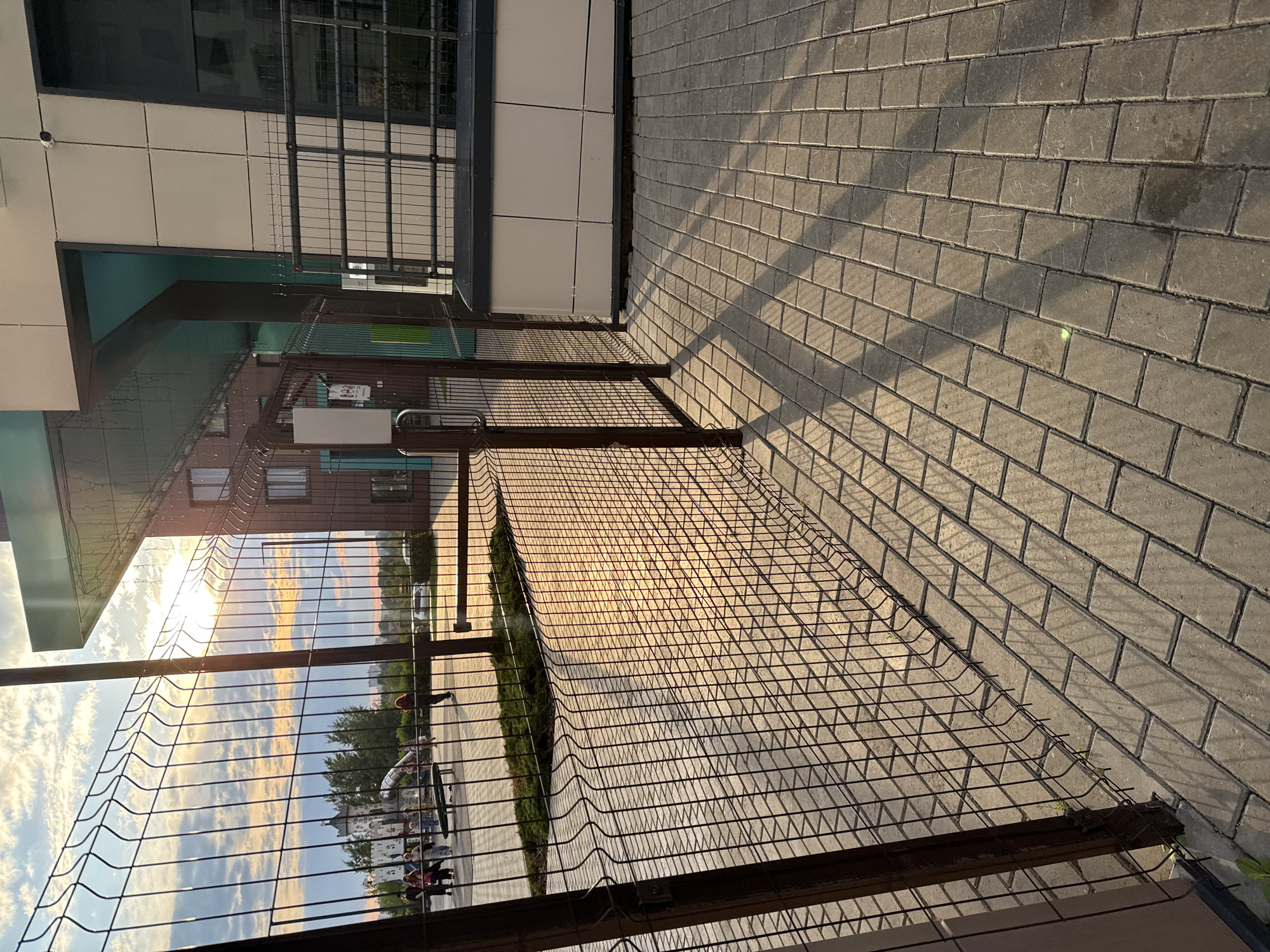 The image shows a fenced-off area of a modern building complex, likely in Perm, Russia, based on the architectural style and the presence of what appears to be a playground. The sun is low in the sky, casting long shadows across the paved walkway and the wire mesh fence. Through the fence, a playground with a castle-like structure and slides can be seen. Several people are present in the playground area, appearing to be adults and children. The building itself has a contemporary design with large windows and varied facade materials. A security camera is visible mounted on the building. The overall scene depicts an outdoor urban environment during the golden hour, possibly a recreational area adjacent to a residential or commercial building.