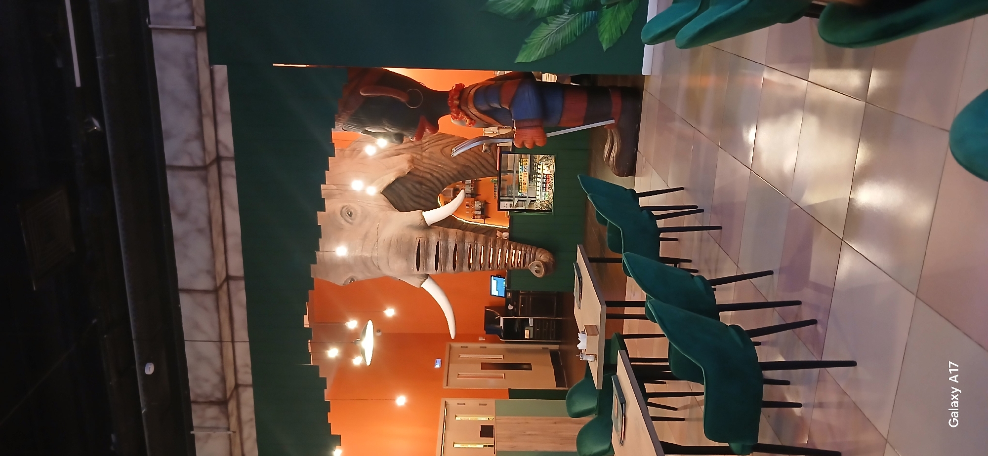 The image depicts the interior of a restaurant in Yekaterinburg, Russia. An elephant head sculpture is prominently displayed on a wall, extending from a central space into the dining area. Above the elephant's head, a human figure sculpture stands, holding what appears to be a spoon or similar utensil. The restaurant features an orange-colored wall with multiple light fixtures, creating a warm ambiance. To the right of the sculptures, tables and green chairs are arranged on a tiled floor. In the background, there's a display case containing food or beverages. The environment suggests a themed dining establishment.