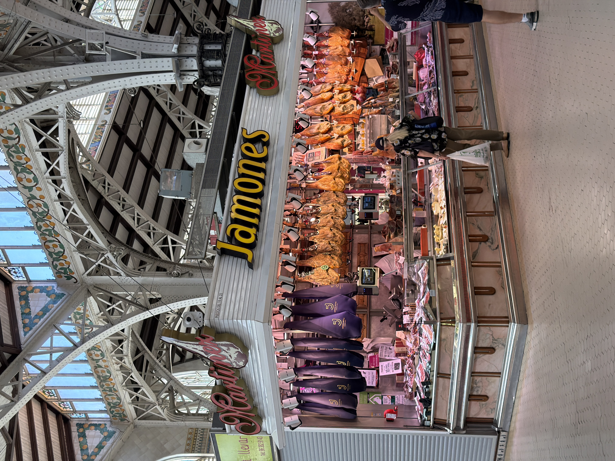 A street market stall displays cured hams and meats under ornate metalwork and stained-glass ceiling in what appears to be a large public building. The stall has large signs that say "Jamones" and "Martinez". Several whole cured hams are hanging from the ceiling of the stall, some in dark purple fabric bags and others exposed to view. Display cases in front of the stall hold various cuts of meat. Two people are visible in front of the stall: one woman in a floral shirt and brown pants holding a white plastic bag, and another person whose back is to the camera wearing a dark patterned shirt and shorts. The floor is tiled with a geometric pattern. The building's architecture features large metal beams and arched windows with colorful decorative elements.