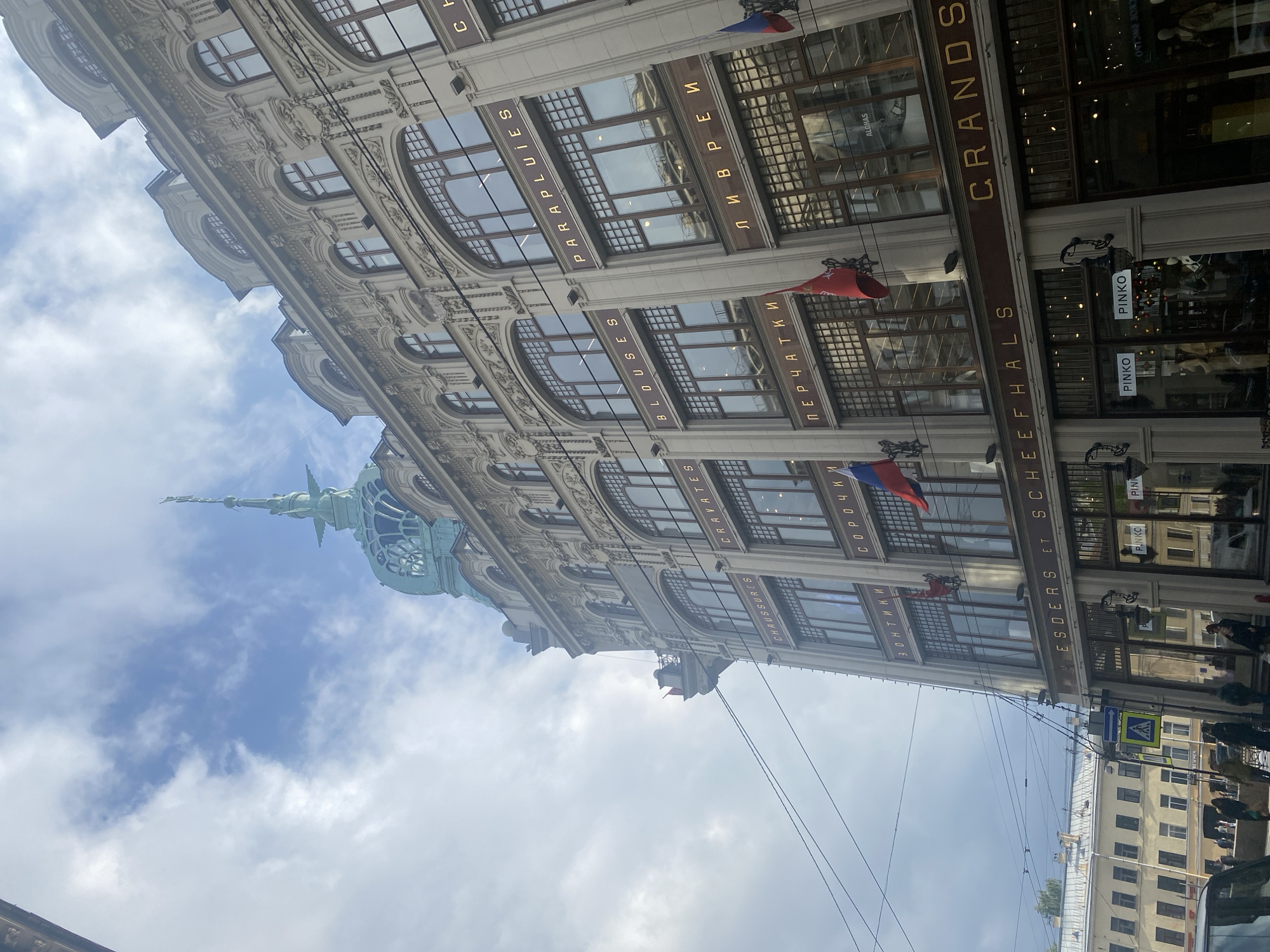 The image shows a street view of the ESDERS ET SCHEEFHALS building in Saint Petersburg, Russia, recognizable by its ornate architecture and the flags adorning its facade. The building houses multiple stores, including PINKO and ALOHAS. Signs in both Cyrillic and Latin alphabets indicate the types of goods sold inside, such as umbrellas, shirts, gloves, and liveries. Power lines are visible against a backdrop of a partly cloudy sky. People are present at street level.