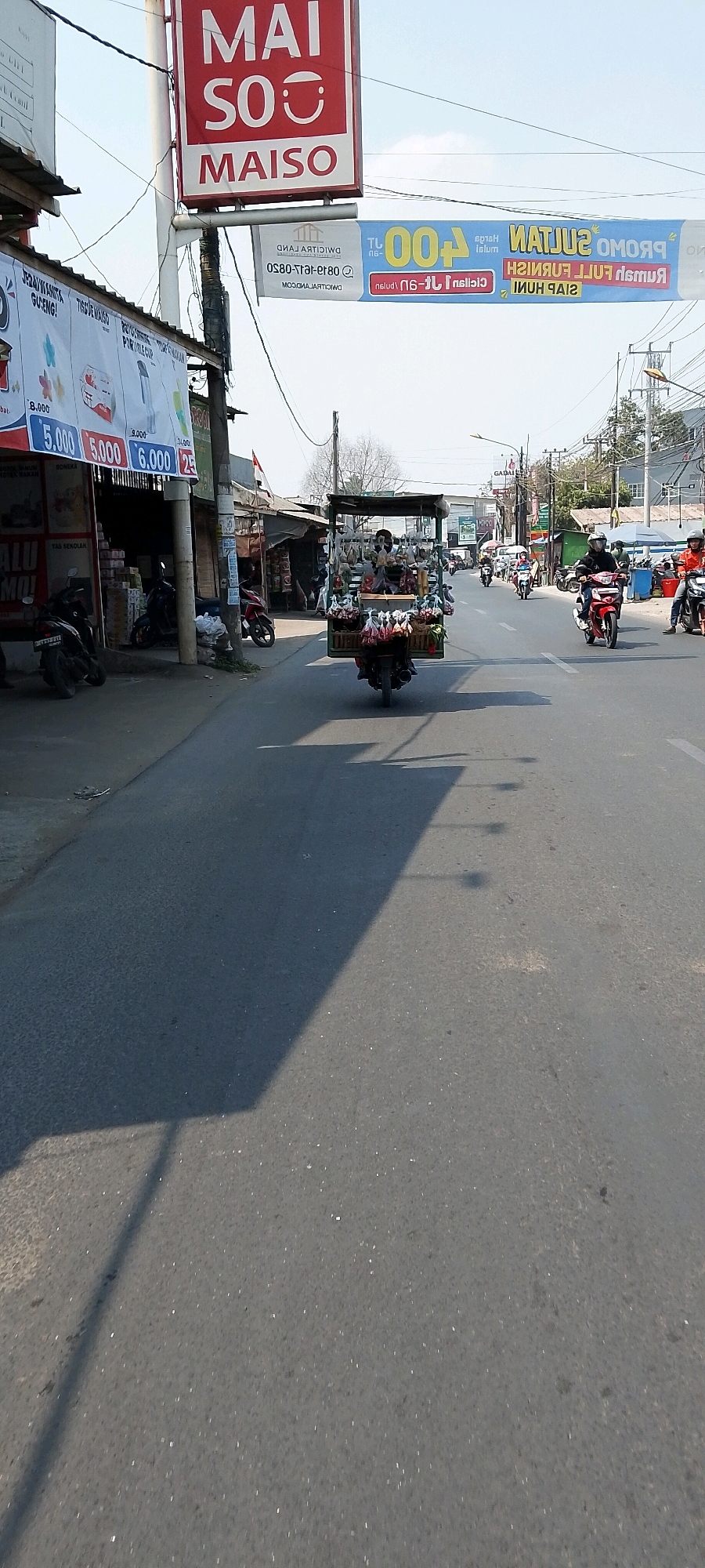 The image depicts a street scene in Babelan, Indonesia, likely on a sunny day, given the clear sky and shadows. In the foreground, a road surface is visible, with a prominent shadow cast by a structure to the left, possibly a building or a large sign. To the left of the road, a row of shops or stalls are visible. One stall has a large red and white sign that reads "MAI SO MAISO." Further down, banners are displayed, including one with blue and yellow colors. A motorcycle carrying a large load of goods, possibly fruits and vegetables, is moving away from the viewer down the center of the road. The motorcycle has a wooden structure on its back, laden with items in plastic bags. Several other motorcycles and a few people are visible further down the street, indicating ongoing traffic and activity. The buildings lining the street appear to be commercial establishments.