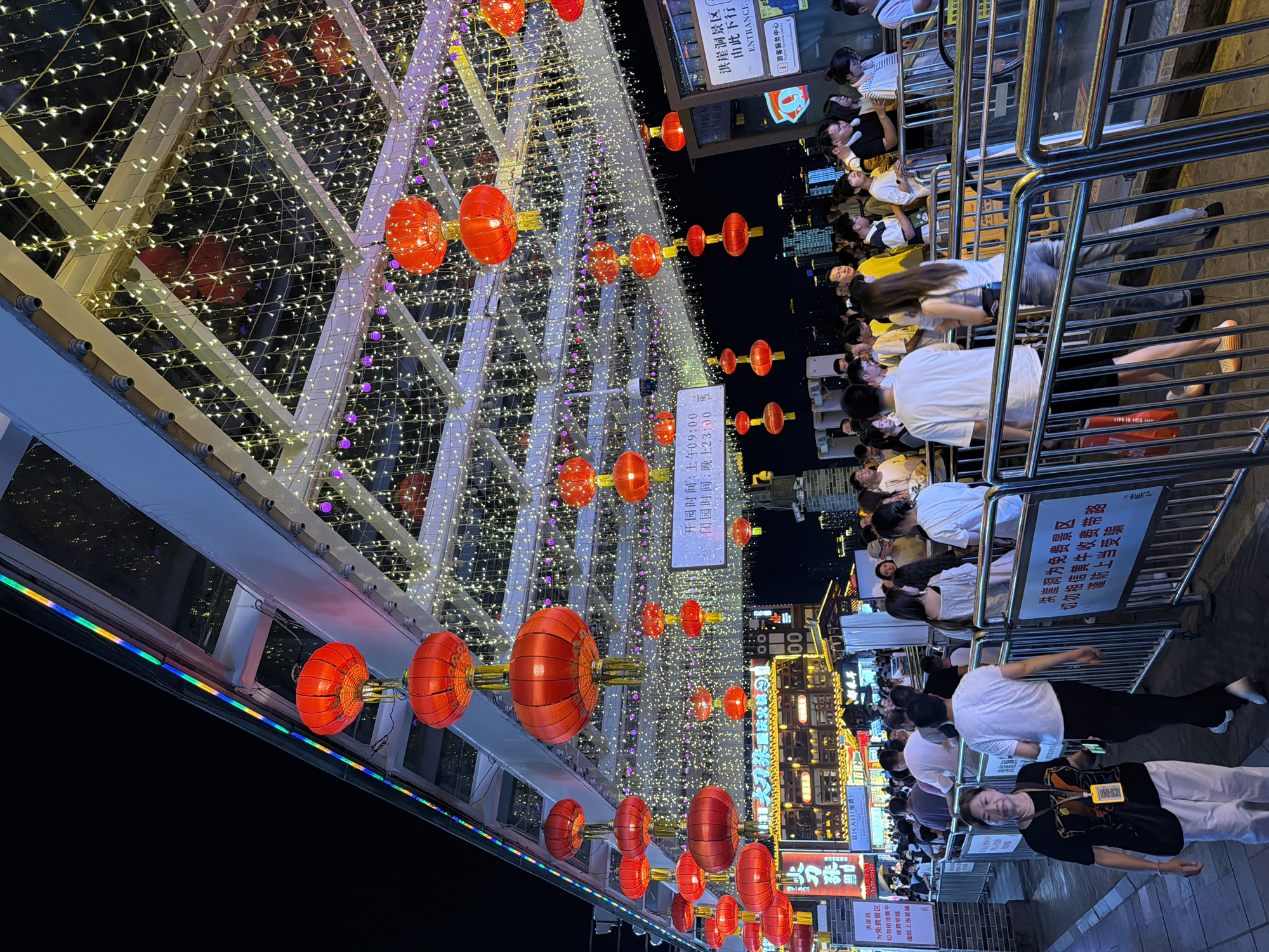 This image captures a bustling night scene in the Yuzhong District of China, specifically at a location adorned with numerous red lanterns and string lights. The scene appears to be an entrance or a thoroughfare, possibly part of a tourist attraction or a lively market. A crowd of people, dressed in casual attire, are visible, some queuing behind metal barriers.
In the foreground, a woman in a black t-shirt and white pants, carrying a bag, is looking towards the camera. Behind her, a man in a white t-shirt and black pants walks away. The barriers are indicative of crowd control, suggesting an organized entry or queuing system.
Above the crowd, the overhead structure is decorated with rows of red lanterns of varying sizes, interspersed with numerous white and purple string lights, creating a festive and vibrant atmosphere. The lights reflect off the translucent ceiling panels, adding to the luminous quality of the scene. In the background, illuminated signs and buildings suggest a commercial or entertainment area.
The overall impression is one of a lively evening, possibly during a festival or a busy tourist period, in a well-decorated urban area of China. The presence of signage in Chinese characters confirms the location.