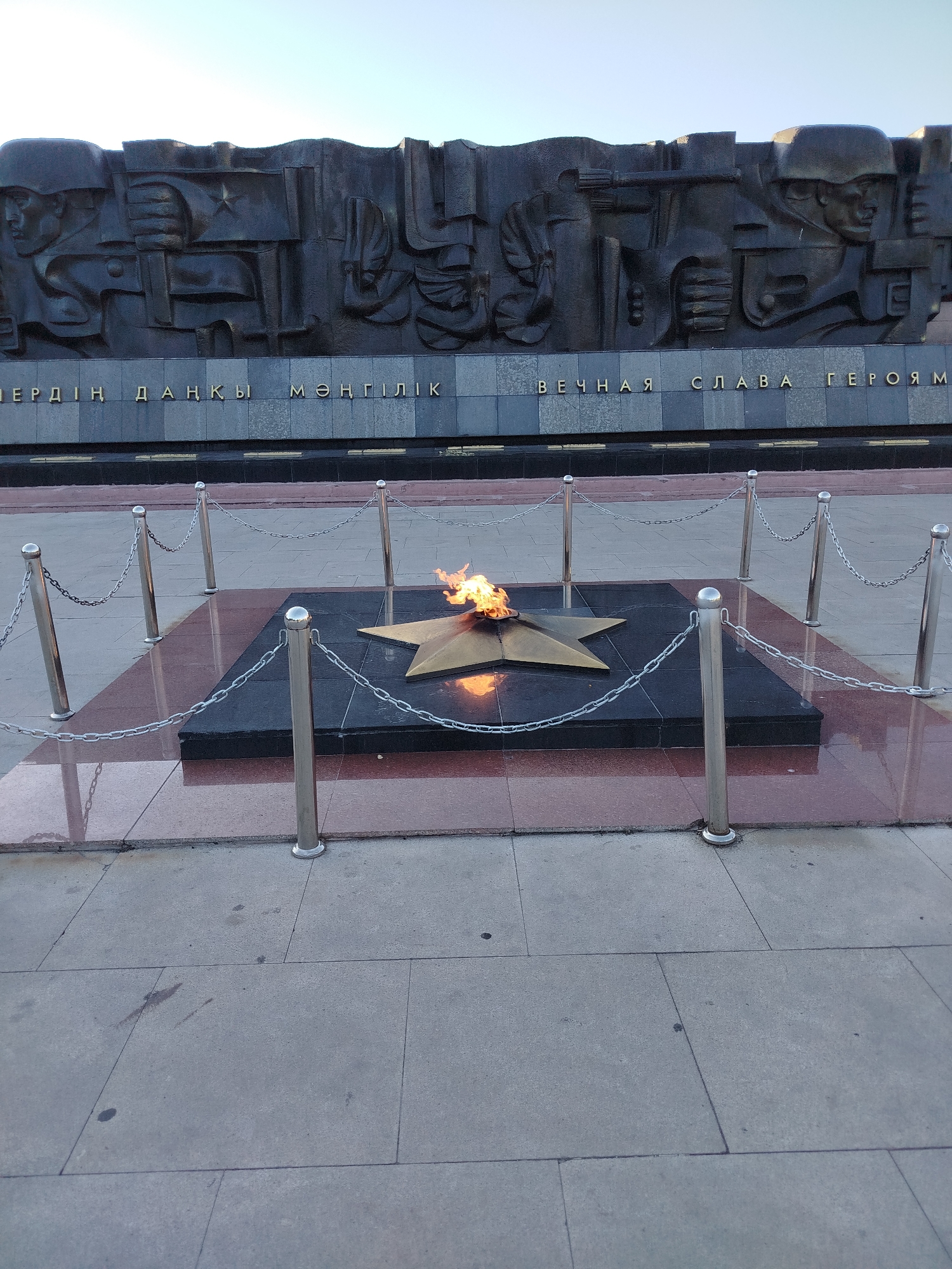 This is a medium-shot, eye-level, outdoor photograph of the Eternal Flame memorial located in Karaganda, Kazakhstan. The memorial features a dark bas-relief frieze depicting soldiers and flags, with inscriptions in both Kazakh and Russian. Below the frieze is a prominent golden five-pointed star with an eternal flame burning at its center, surrounded by a polished black granite base and a red granite border. The memorial is enclosed by a metal stanchion and chain barrier. The surrounding ground is paved with large, light-colored square tiles. The sky above is a pale, clear blue, indicating daytime. The image captures a solemn and commemorative atmosphere.
