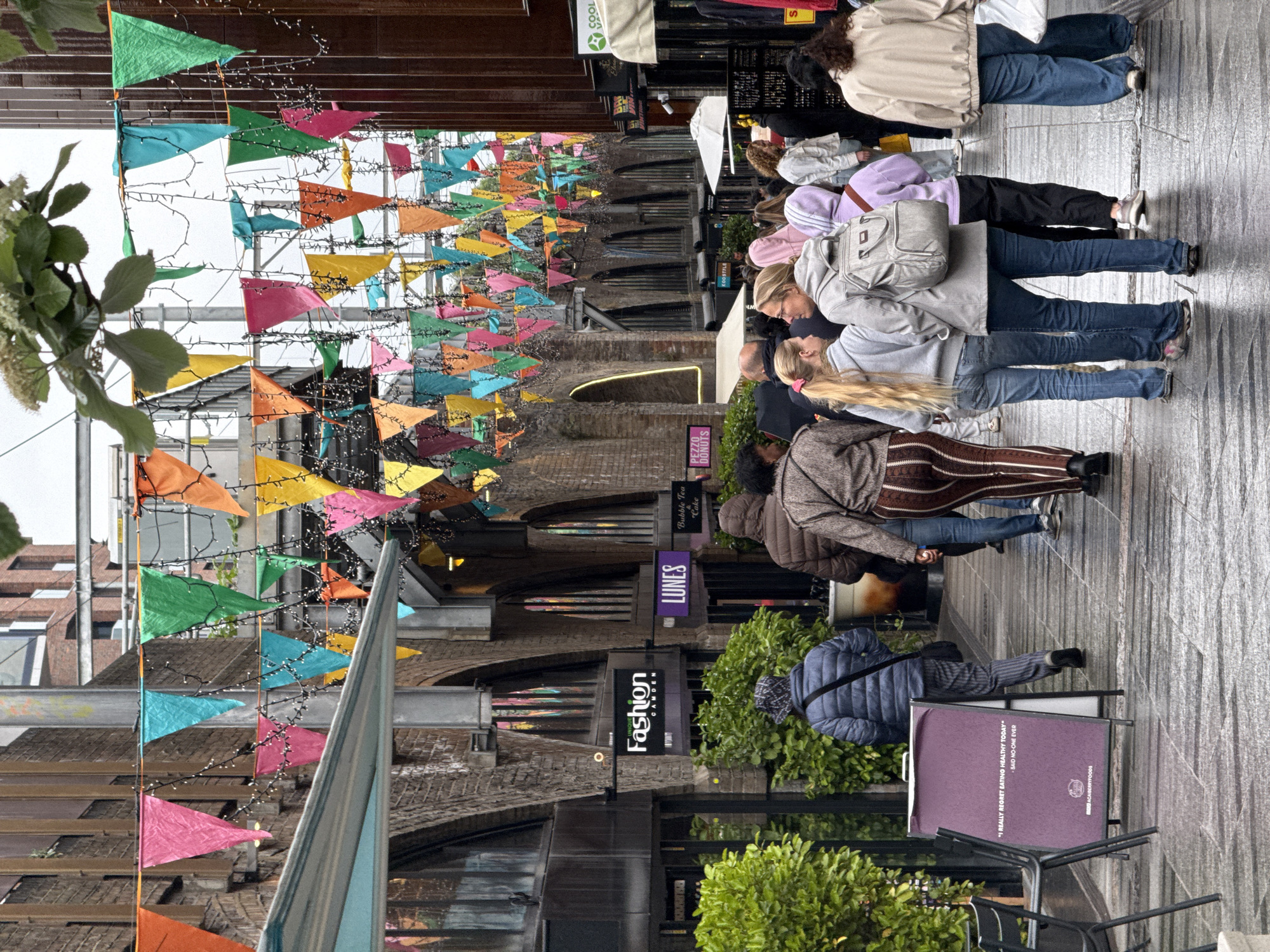 A street in London, United Kingdom is filled with a diverse crowd of people walking. Above, colorful triangular flags are strung across the alleyway, adding a festive touch to the urban scene. Buildings line both sides of the street, exhibiting a blend of architectural styles. A sign reads "Fashion Camden" and "Lunes." The ground is paved with a reflective surface. A poster is on a stand.