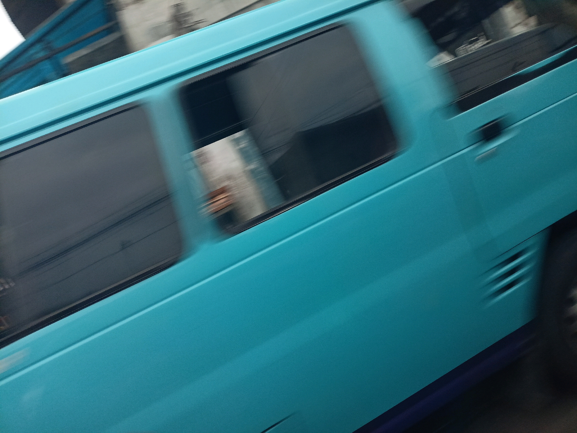 The image shows a side view of a turquoise-colored vehicle, possibly a minivan, in motion. The vehicle has tinted windows, and the motion blur suggests it is moving at a considerable speed. The background is also blurred, indicating movement. On the side of the vehicle, there is a door handle and ventilation slats. The overall scene is captured from a low angle, looking upwards towards the vehicle. It is not possible to determine the specific location within Kebonlegi, Indonesia, from the provided image.