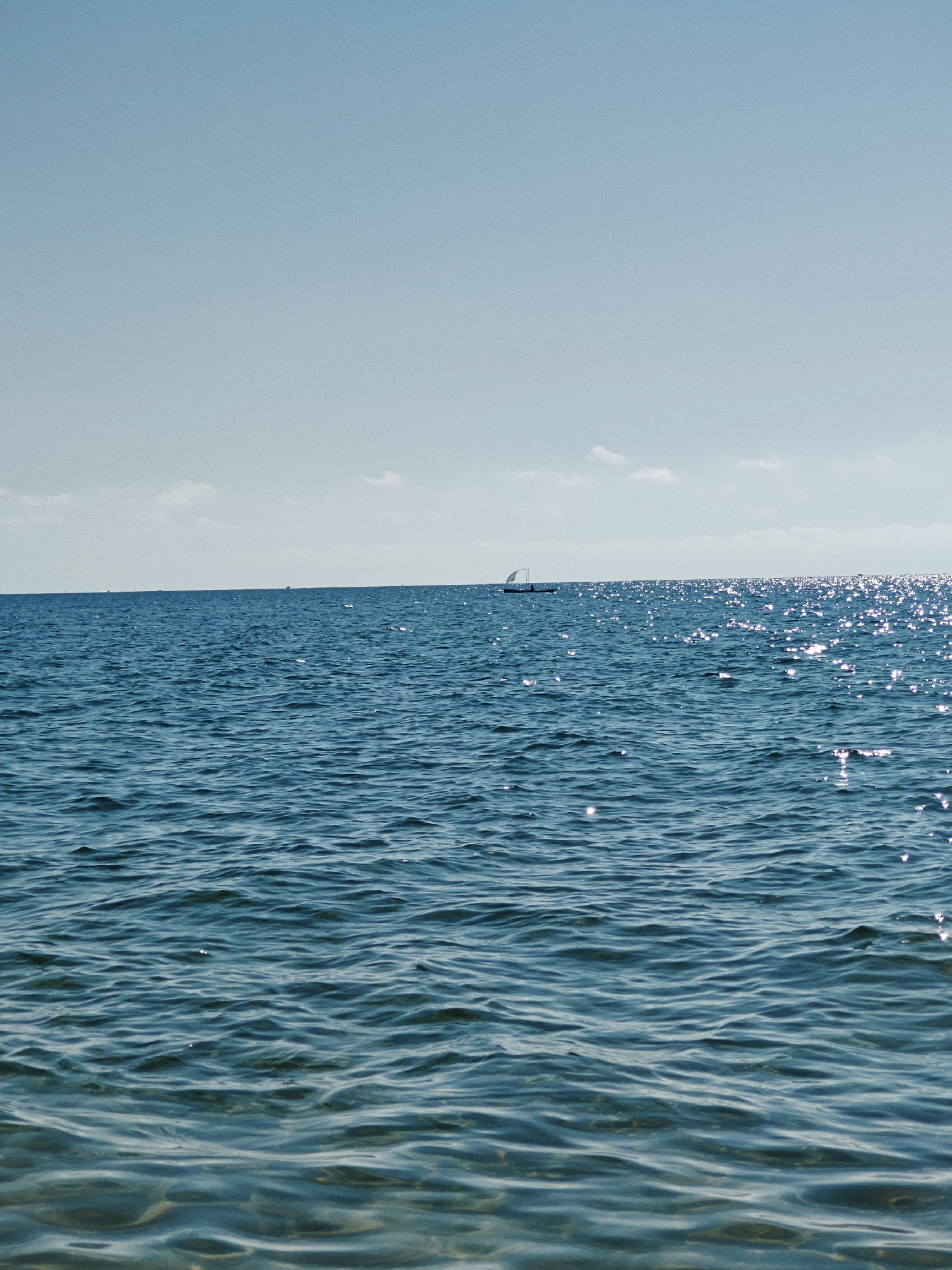 A serene seascape under a clear blue sky features a vast expanse of deep blue ocean water. The water's surface is rippled, with sunlight glinting off the waves, creating a shimmering effect. In the distance, a small sailboat with a single white sail navigates the water. The horizon line is sharp, separating the ocean from the pale blue sky, where a few wispy white clouds are scattered. The overall scene evokes a sense of tranquility and open space.