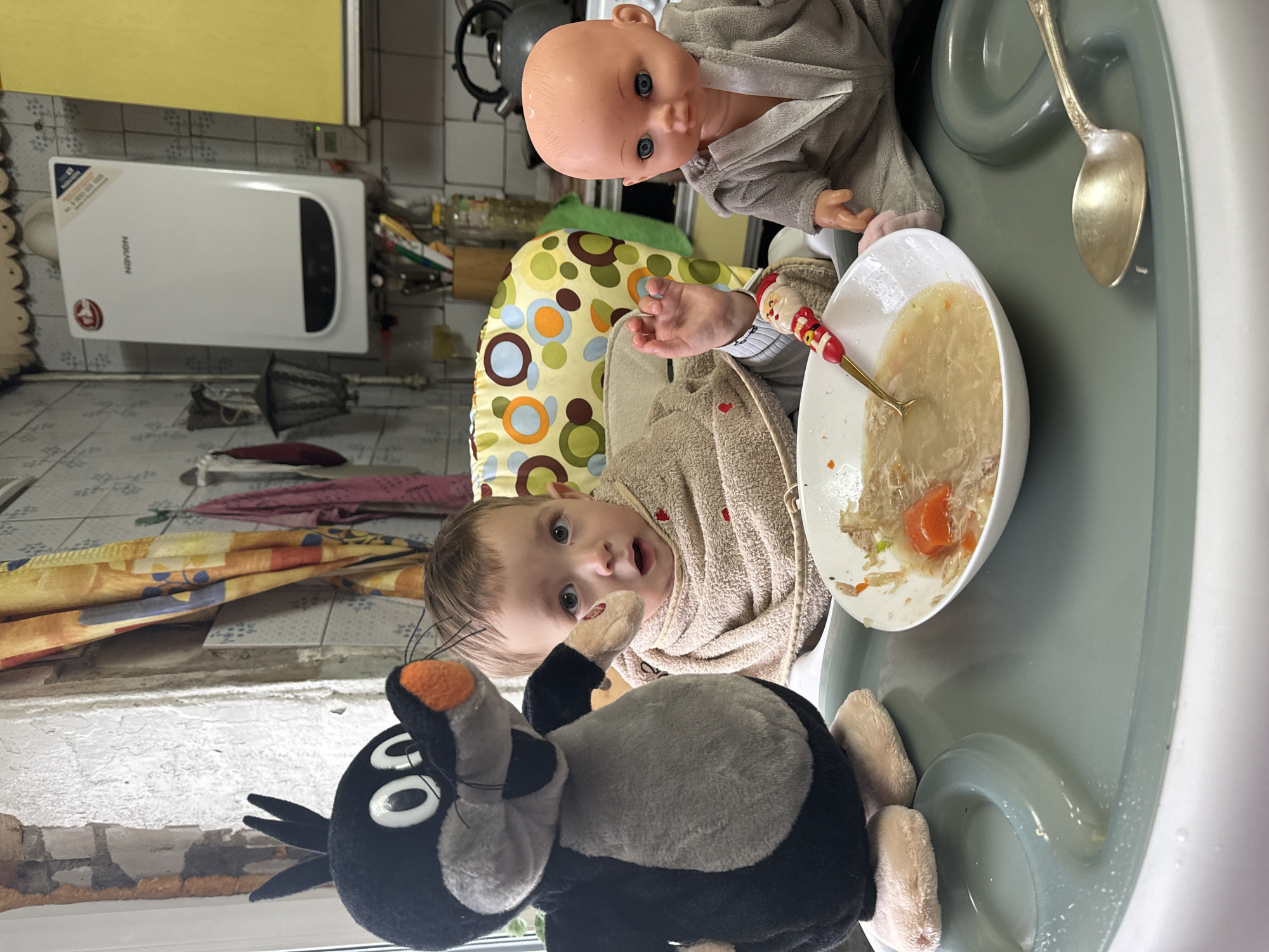 A vertically-oriented indoor photograph depicts a child sitting in a highchair with a bowl of soup, accompanied by a doll and a stuffed animal.
The child, situated in the lower-center of the frame, is wearing a light brown robe. They are looking directly at the camera. A black and grey stuffed animal is positioned near the lower edge of the image, slightly to the left. Above and to the right of the child, a doll is also seated in the highchair, wearing a matching robe.
A white bowl of soup, containing discernible pieces of carrot and meat, sits on the highchair tray, partially eaten. A spoon with a Santa Claus design is also in the bowl. A second, antique-style silver spoon is lying on the right edge of the tray.
In the background, a white water heater is visible on the wall, which is covered in light colored tiles. A metal rack with various utensils and kitchen items hangs to the right of the water heater. A patterned yellow and green chair cushion is behind the child. The room lighting seems to be a combination of natural light coming from the window in the lower left and artificial light from an overhead source.
The location in the city of Unknown, Unknown.