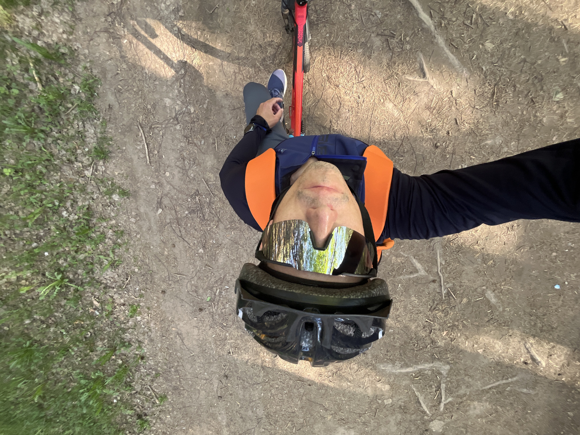 A man on a red mountain bike is positioned on a dirt path in a wooded area. He is wearing a black helmet and mirrored sunglasses. The man is wearing a navy blue and orange vest over a navy blue long-sleeved shirt. He is wearing gray pants and a blue shoe on his left foot. The ground is dirt and scattered with twigs and dry leaves and some green grass.
The exact location in the city of Chaikovsky, Russia, cannot be determined from the given information.